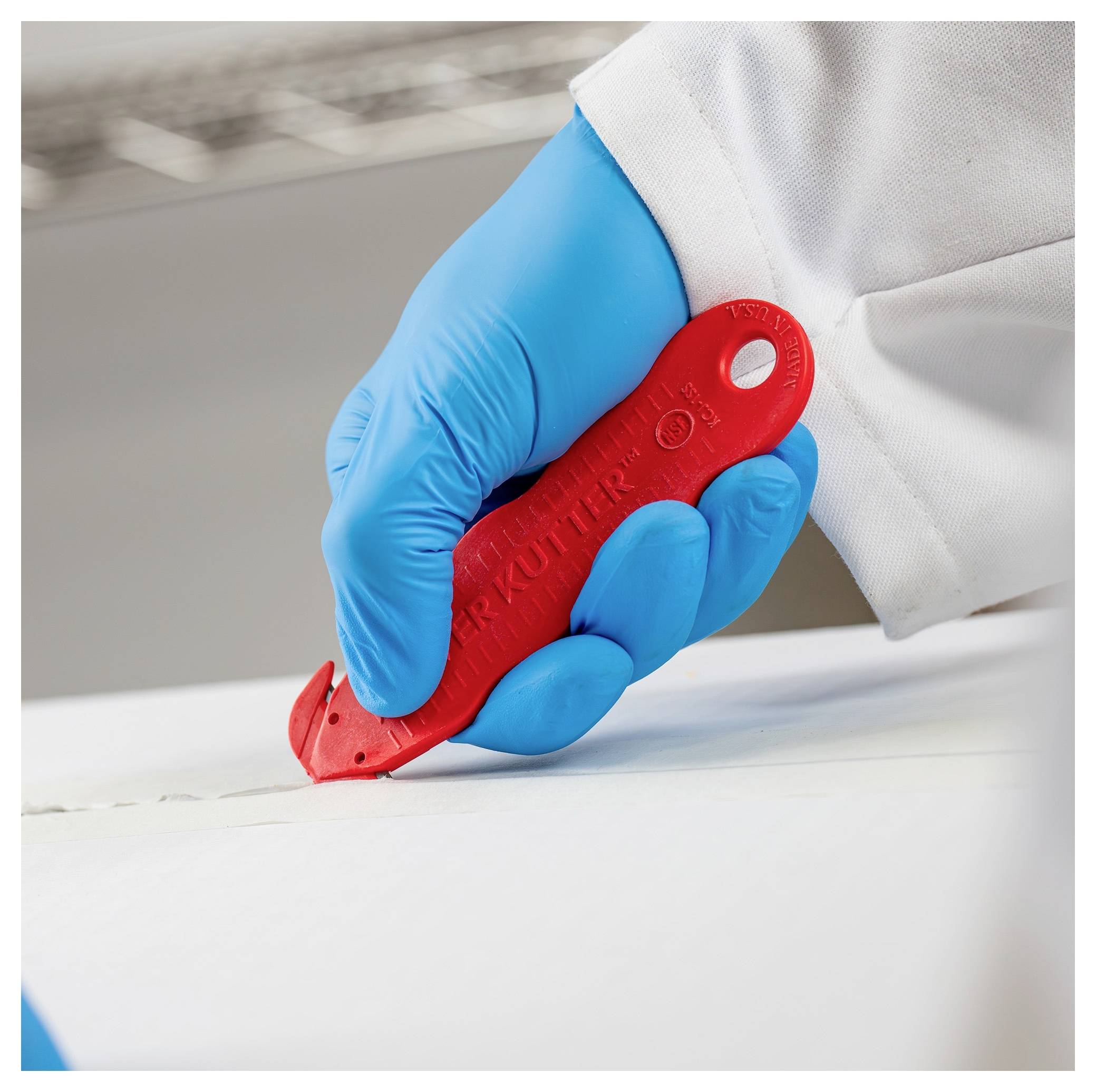 Gloved hand using a red utility knife to cut a white material on a work surface.