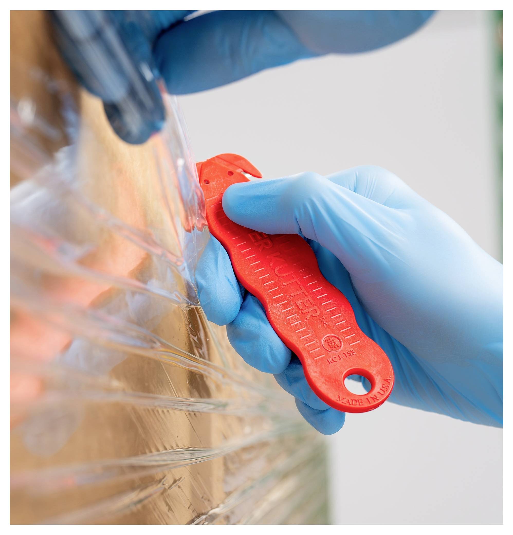 Gloved hands use a red utility knife to cut clear plastic wrap from a cardboard shipping pallet or box.