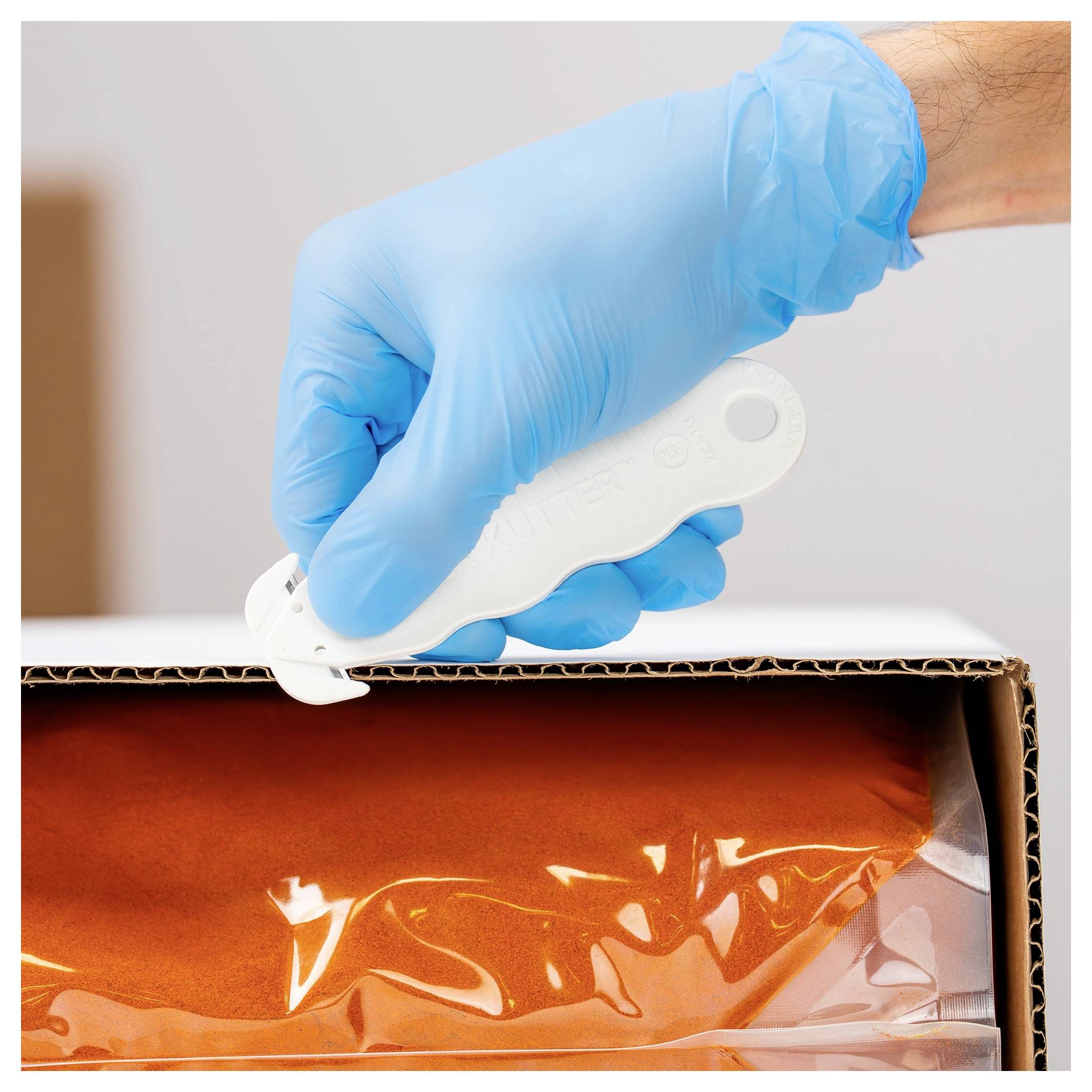 Blue-gloved hand uses a white box cutter to open a cardboard package containing an orange plastic-wrapped item.<br>