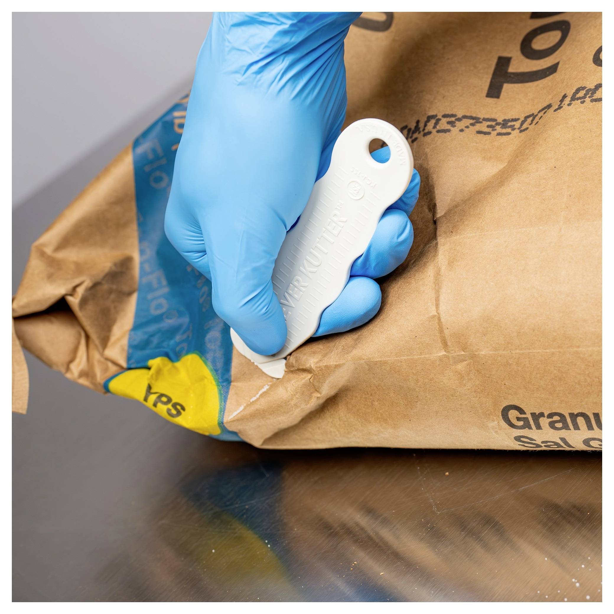 Gloved hand uses a white tearing tool to open a brown paper bag or package on a metal surface.<br>