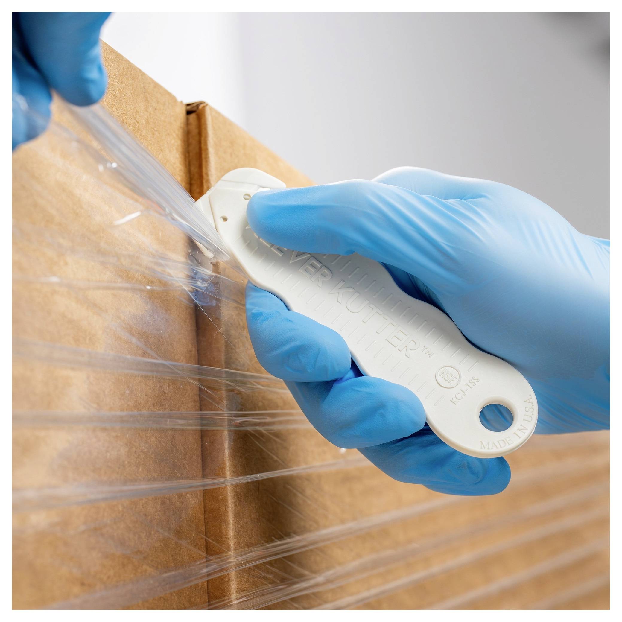 Gloved hands use a box cutter to slice clear plastic wrap from a cardboard box.