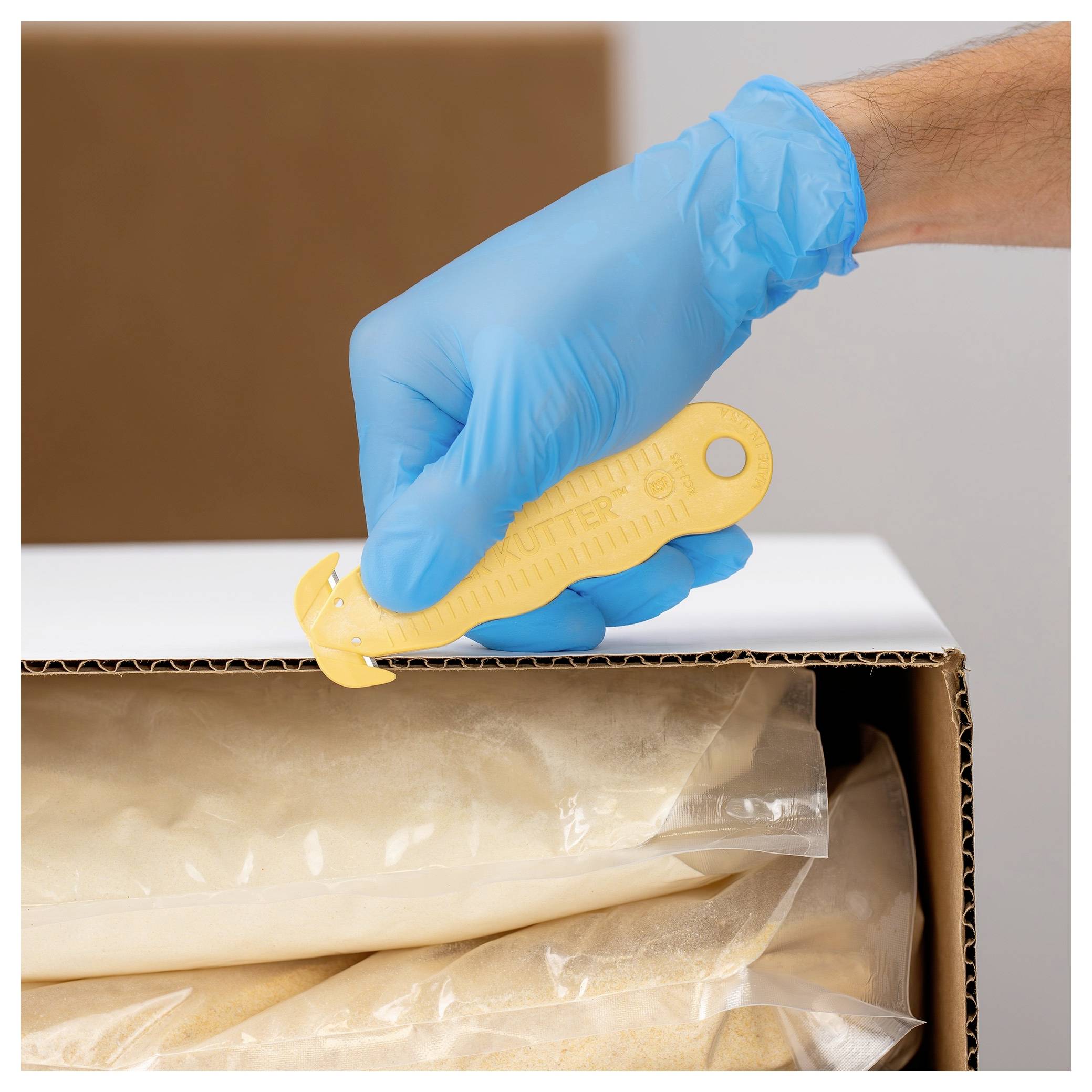 Blue-gloved hand using a utility knife to cut open a cardboard box containing wrapped items<br>