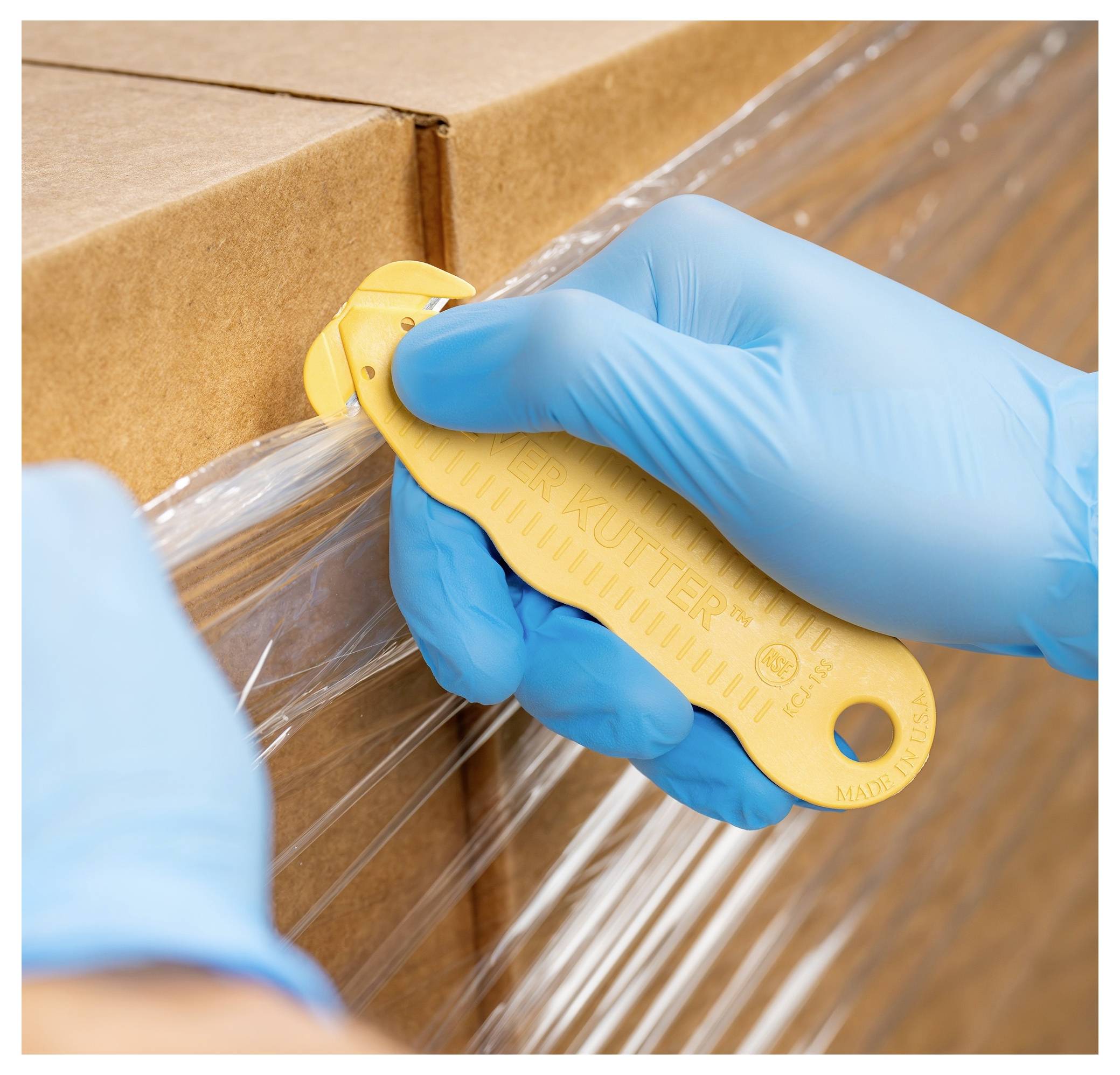 Gloved hands use a box cutter to cut plastic wrap on a cardboard shipping box.