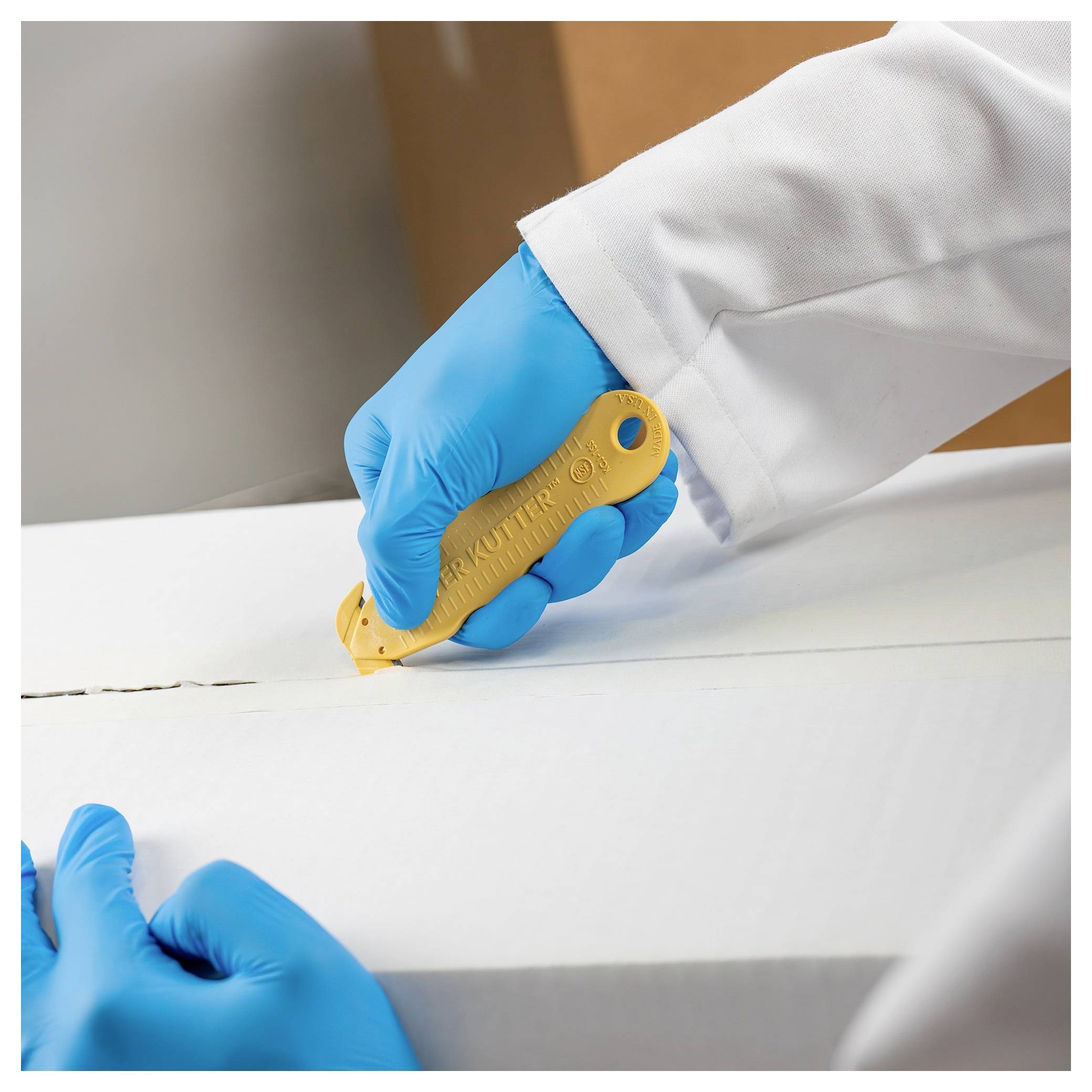 Gloved hands use a utility knife to cut a seam in a white packaging box or foam sheet.
