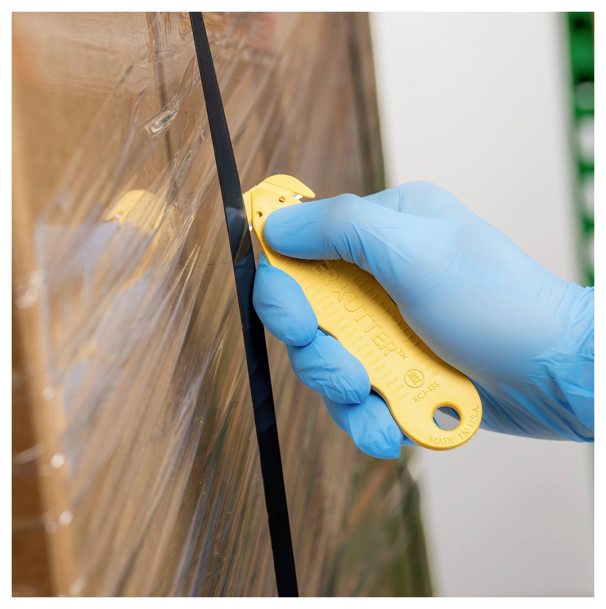Gloved hand cutting clear plastic wrap with a yellow utility knife on a wrapped pallet box.