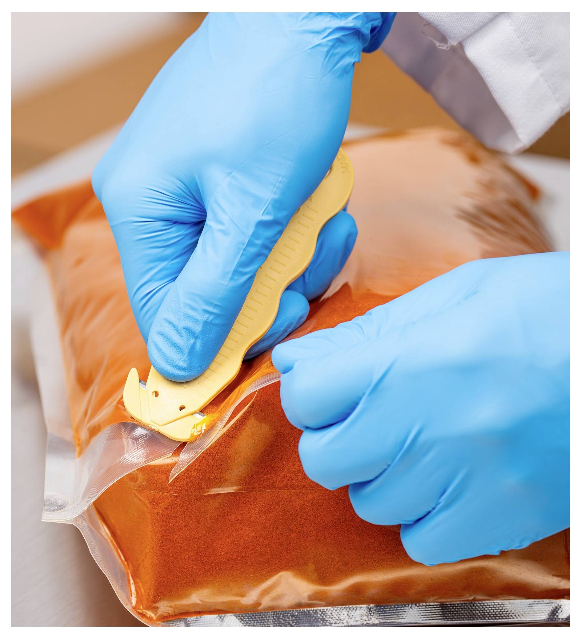Gloved hands use a sealing tool to close a clear plastic bag filled with orange liquid or substance in a lab or food-prep setting.