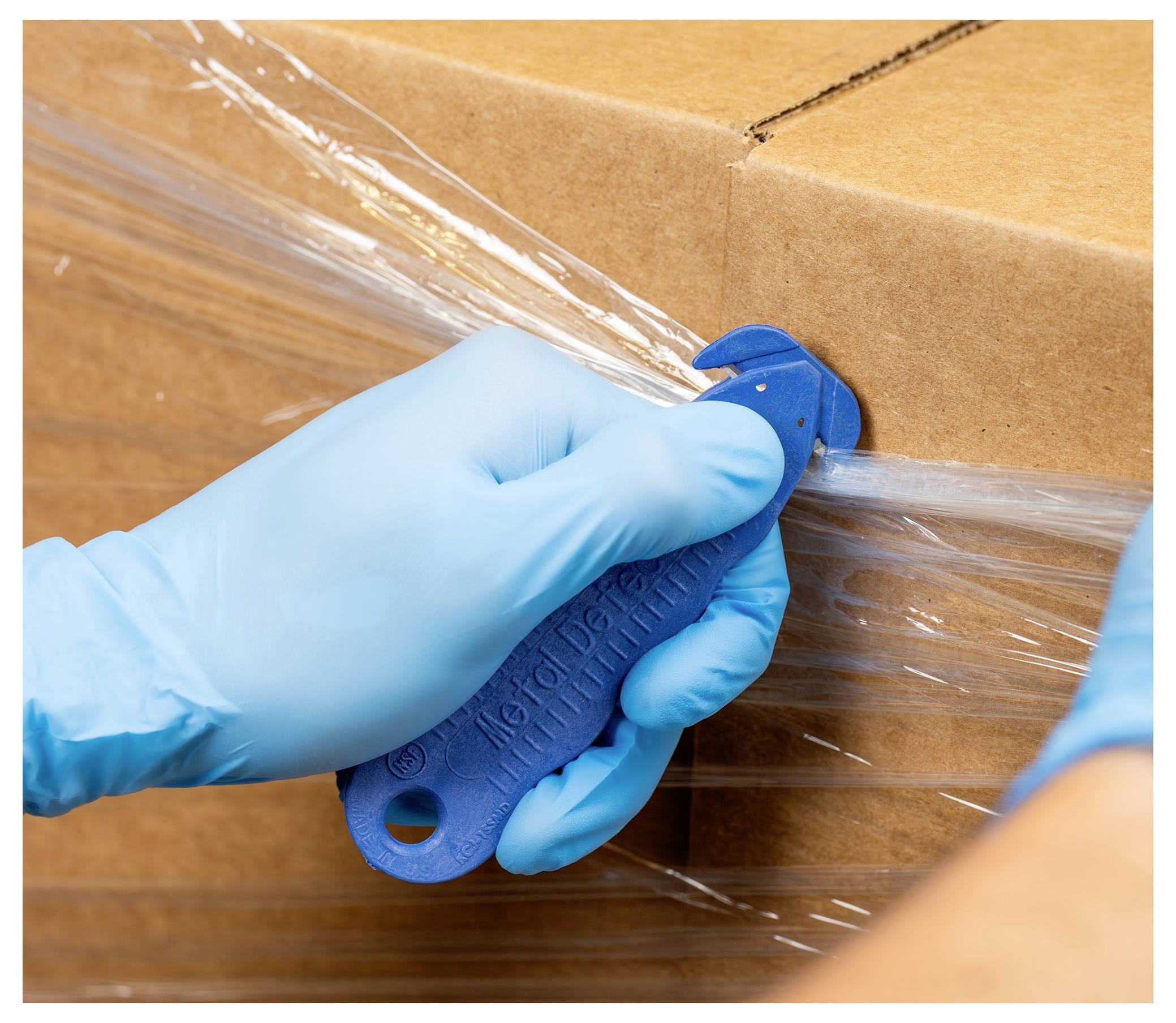 Blue-gloved hand uses a blue box cutter to slice plastic wrap around a cardboard shipping box.<br>