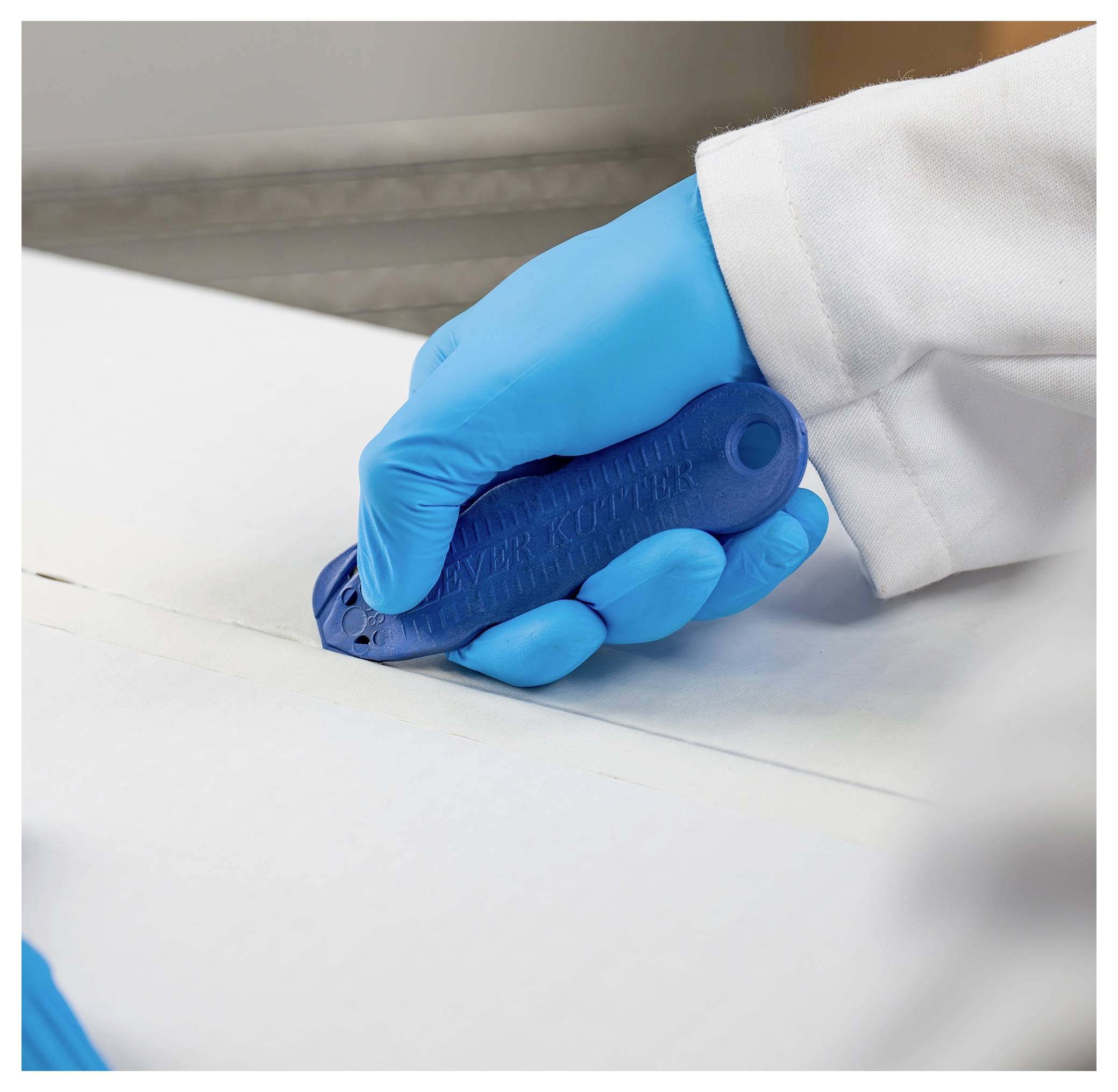 Close-up of a gloved hand holding a blue rotary cutting tool against white fabric, likely cutting material in a work setting.