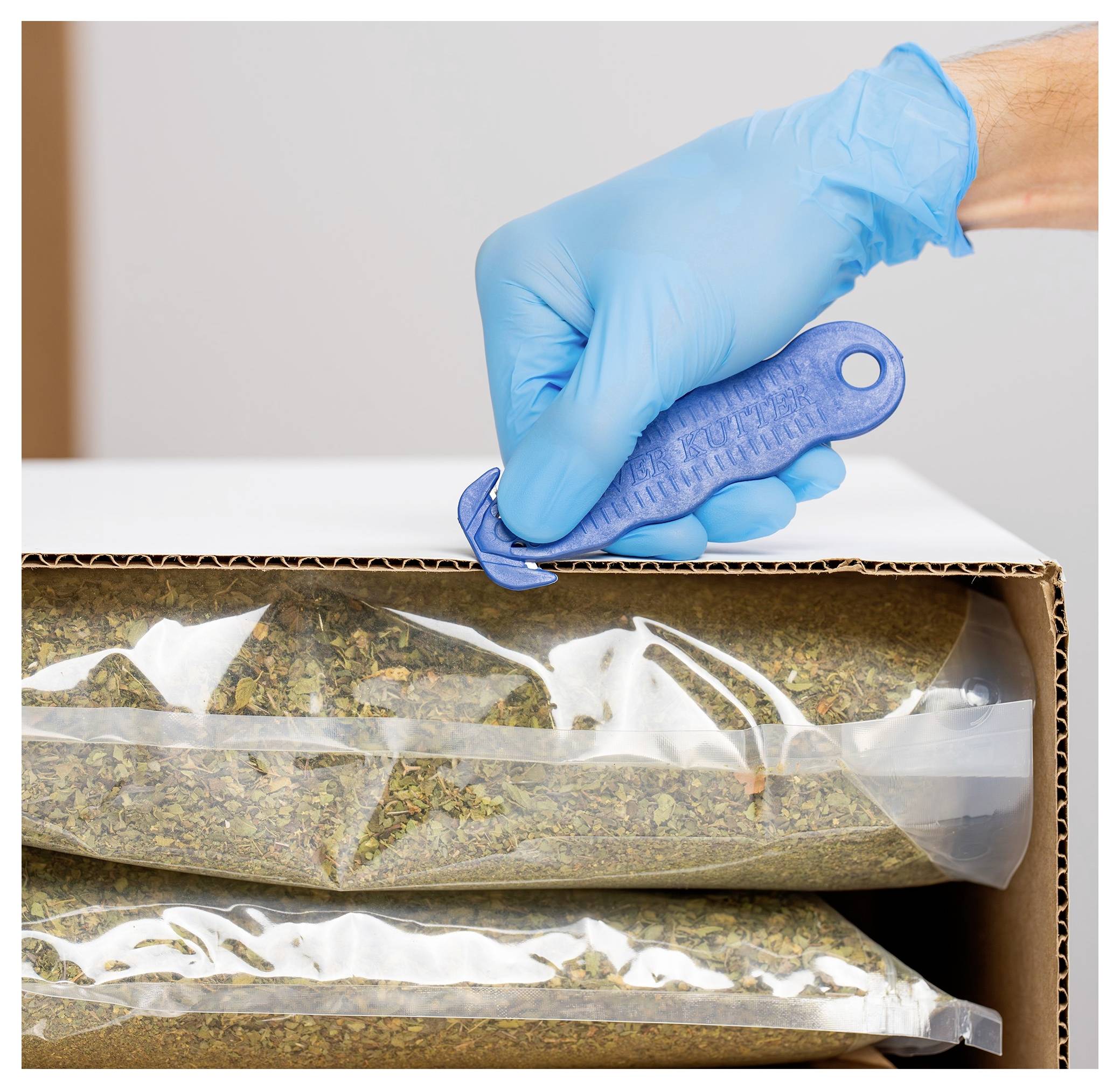 Gloved hand sealing a cardboard box of dried plant material in clear plastic bags using a blue clip.