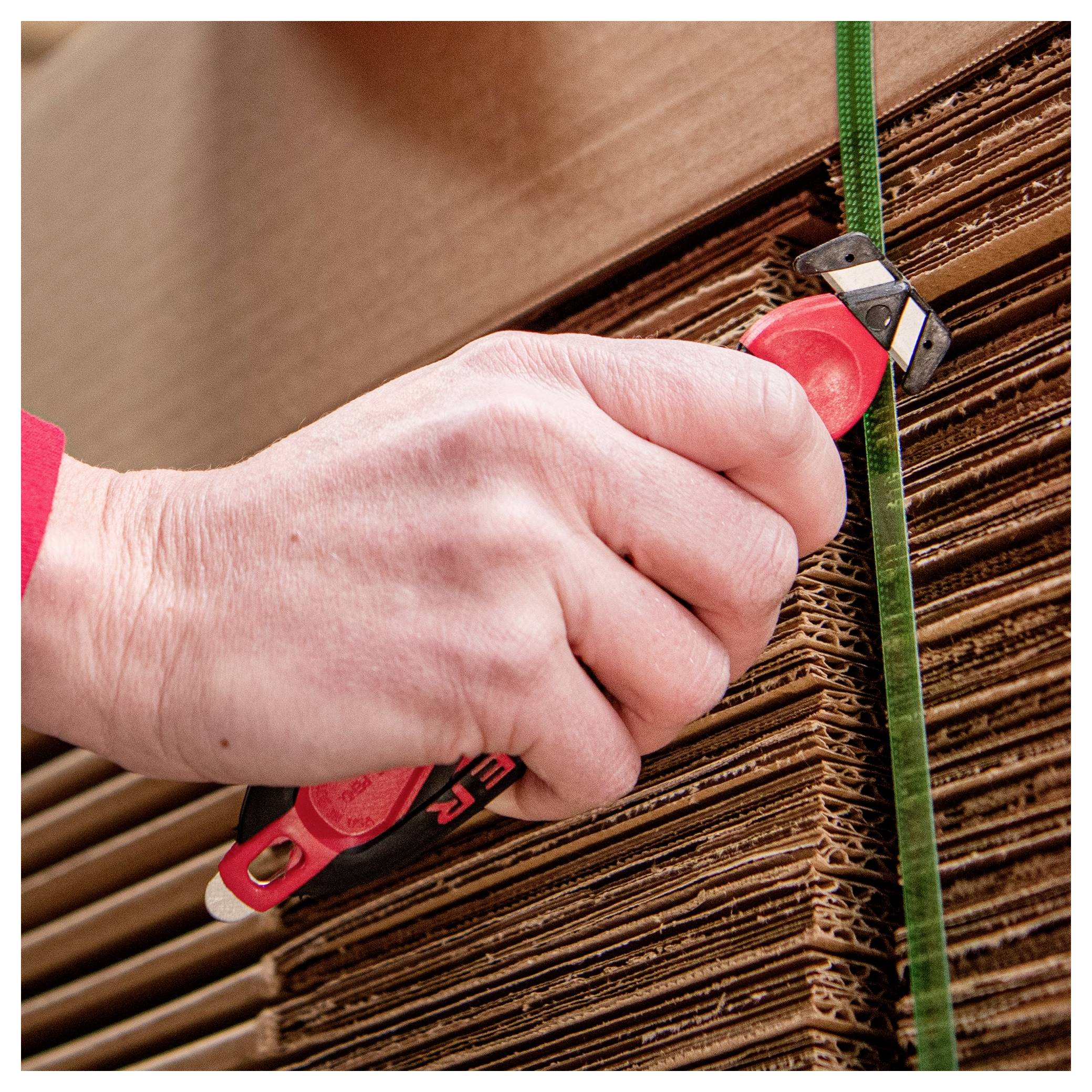 Hand using a red utility knife to cut a green strap securing stacked cardboard sheets.