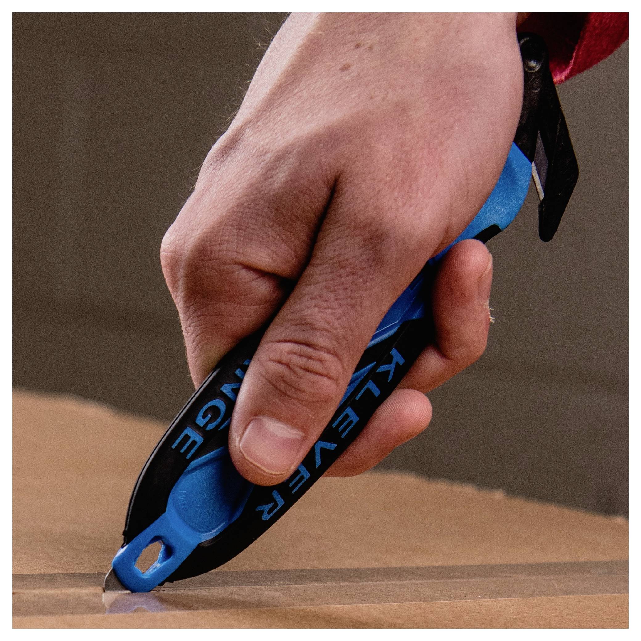 Close-up of a hand using a blue utility knife to cut cardboard on a table.