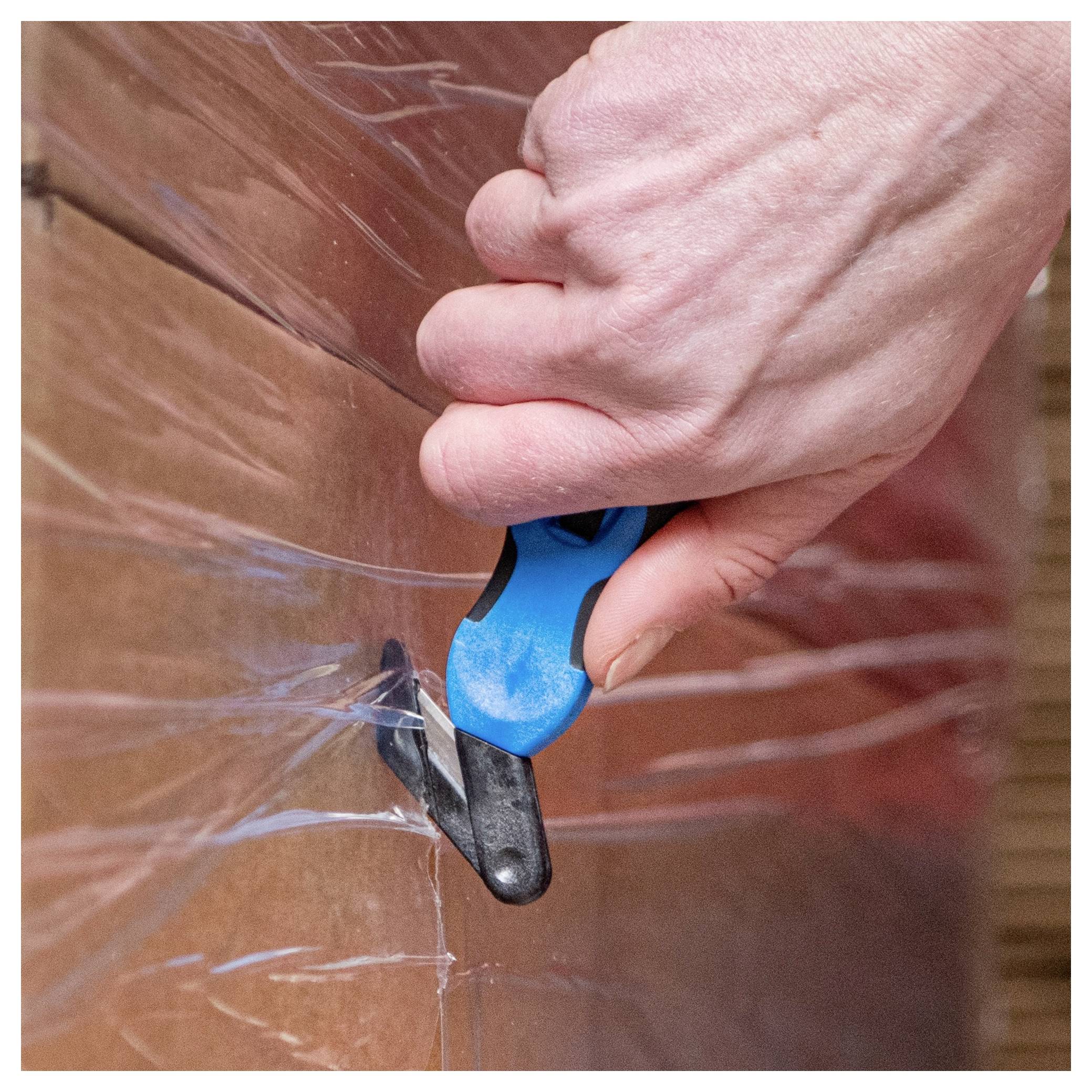 Hand using a blue utility cutter to cut through clear plastic wrap over a large cardboard box.