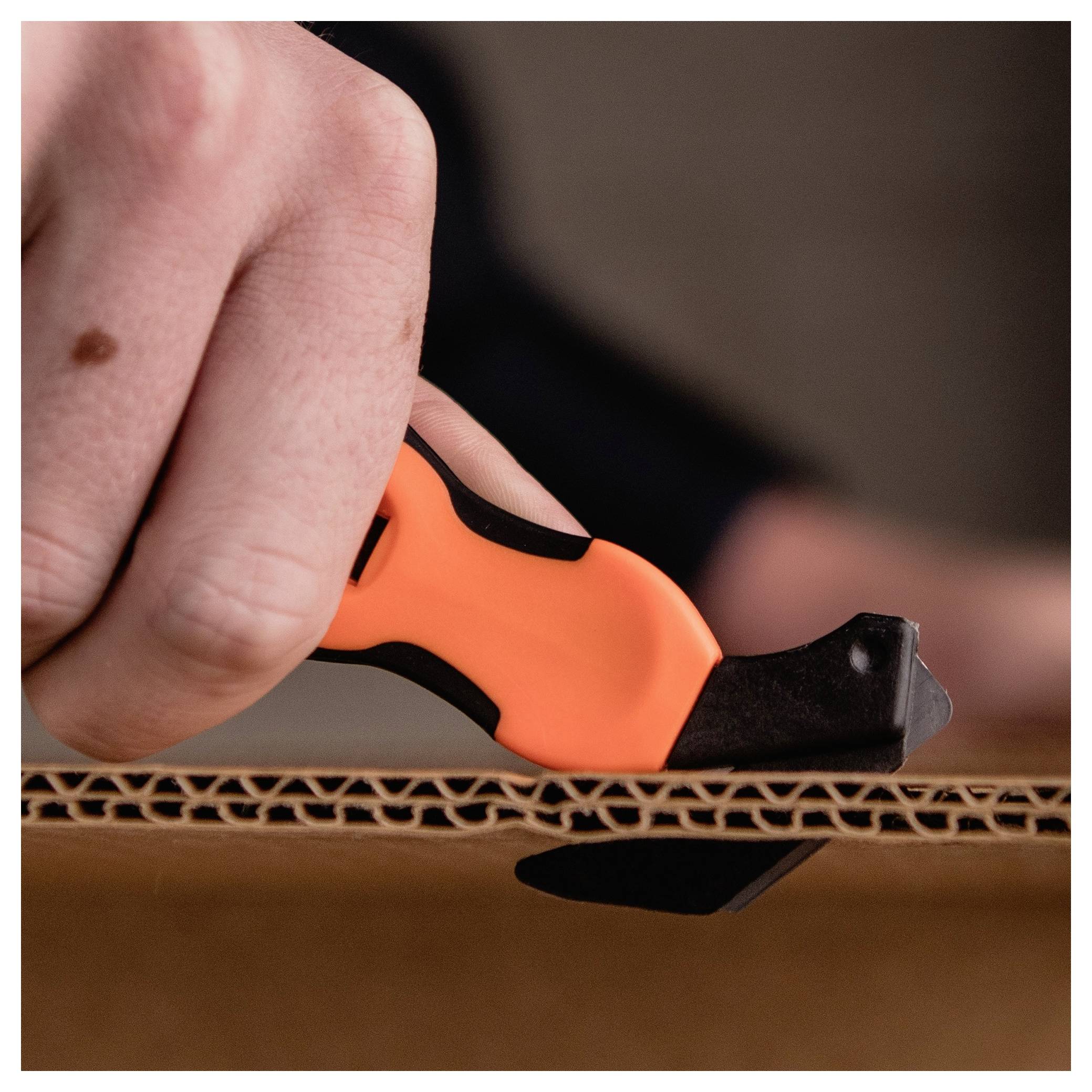 A hand uses an orange utility knife to cut through corrugated cardboard.<br>