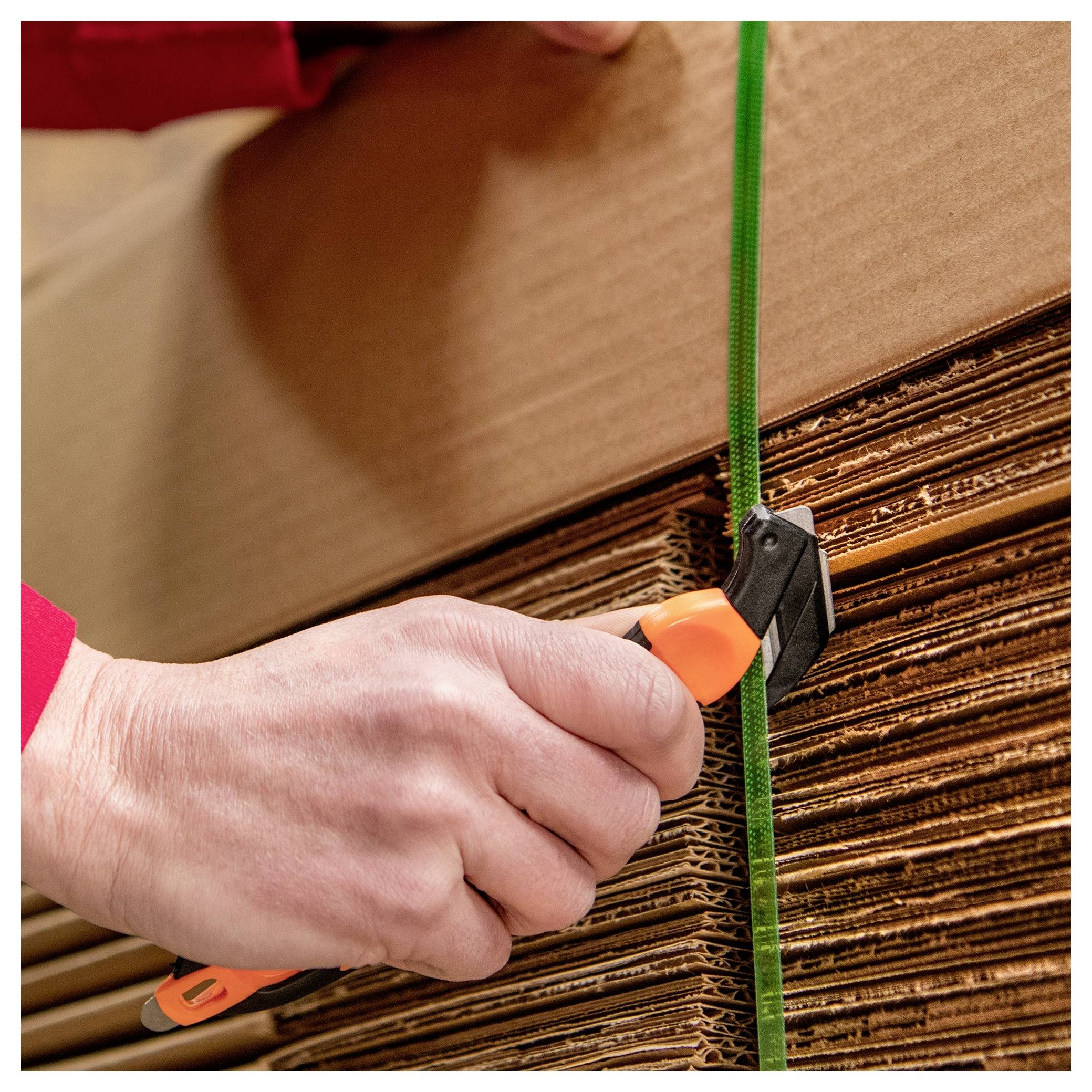 Hand using a strap cutter to cut a green band securing stacked cardboard sheets or boxes.