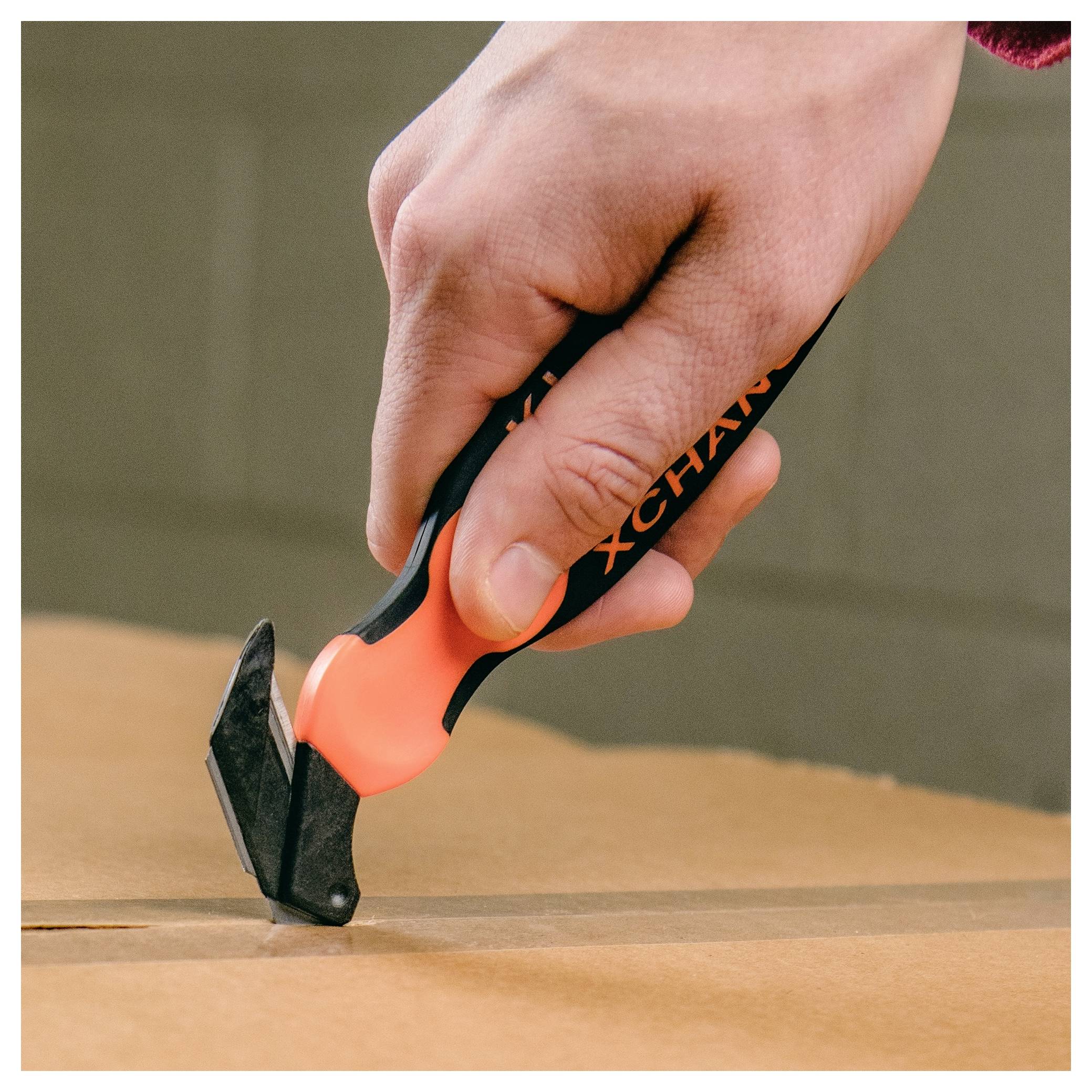 Close-up of a hand using an orange-and-black utility cutter to slice open a cardboard box.<br>