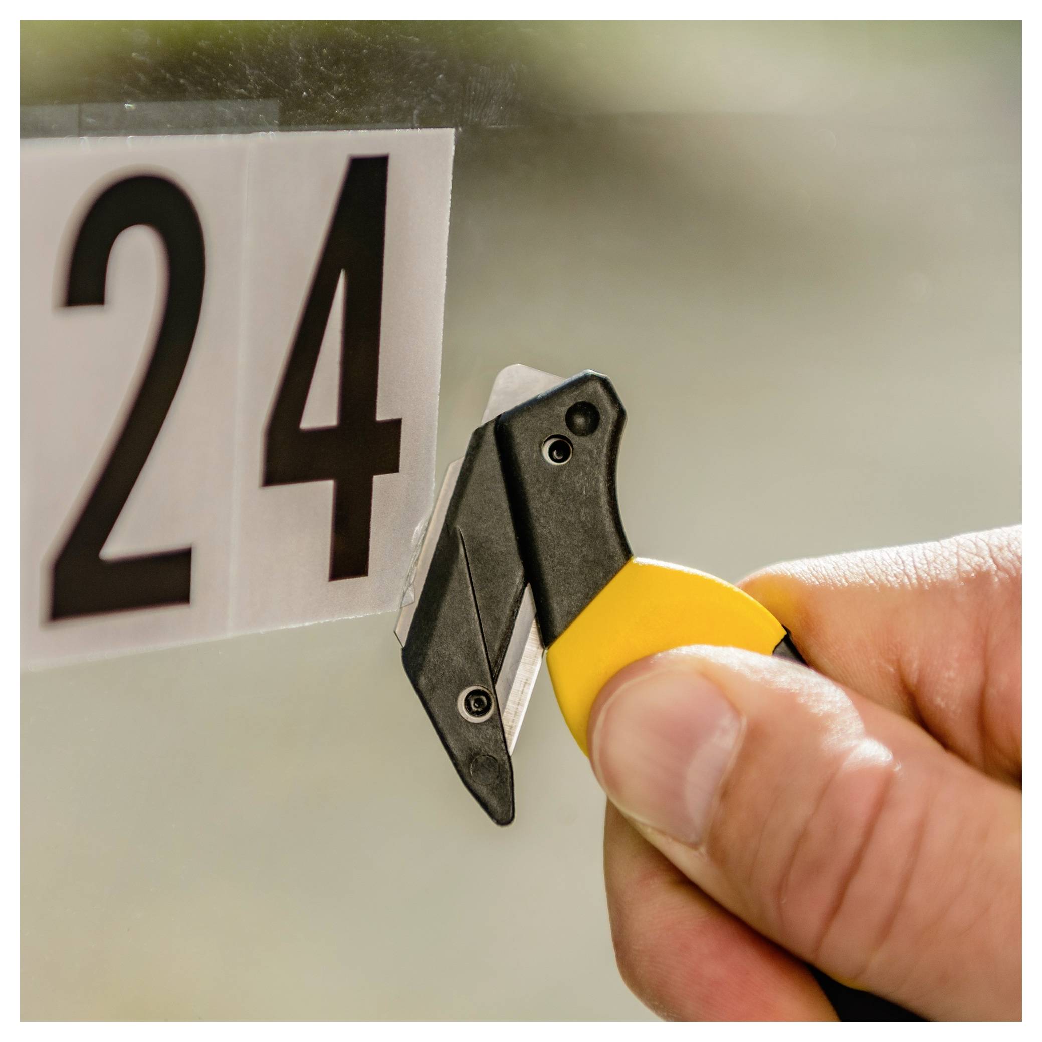 Hand holding a scraper blade near a window or glass pane labeled '24'.