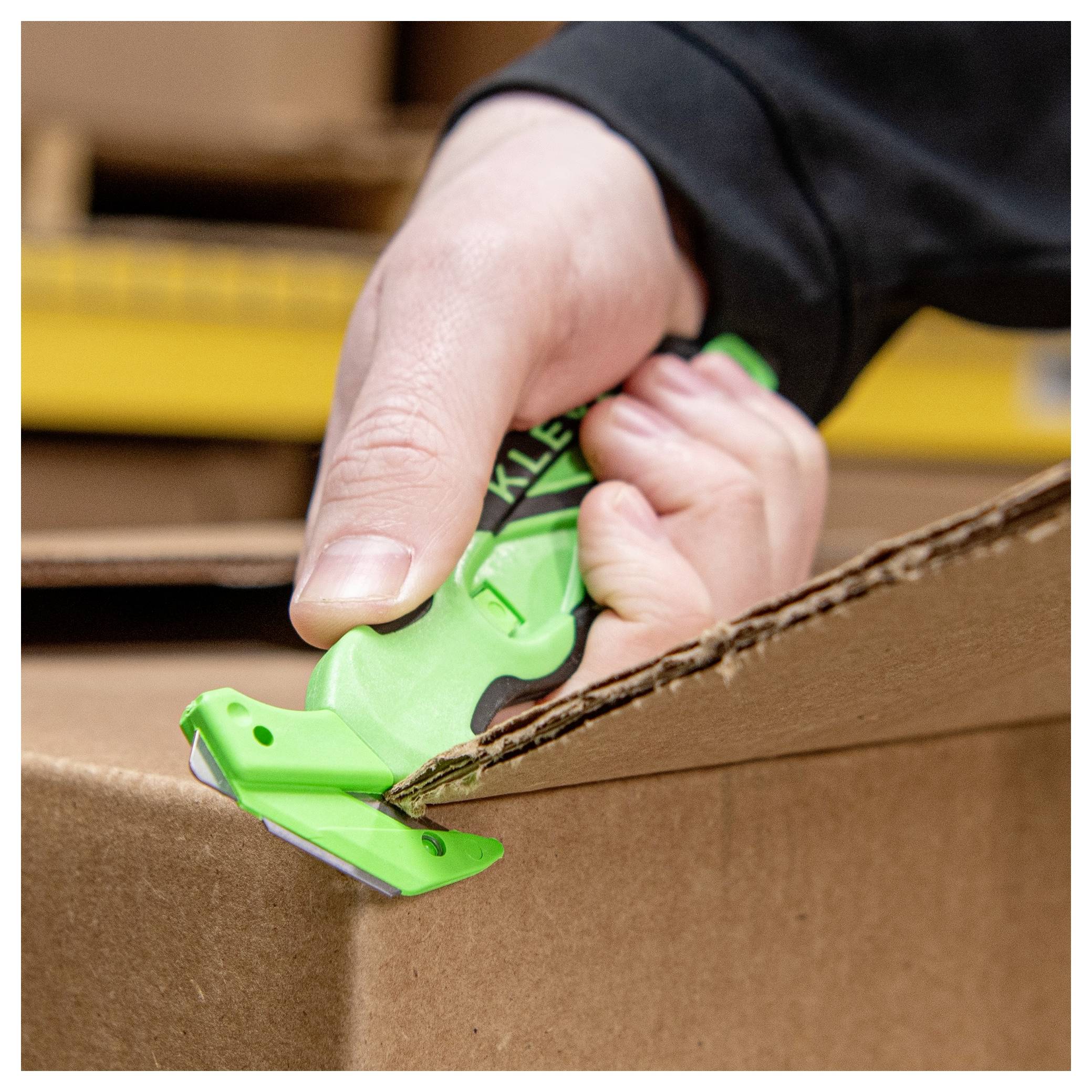 Hand using a green box cutter to open a cardboard shipping box.<br>