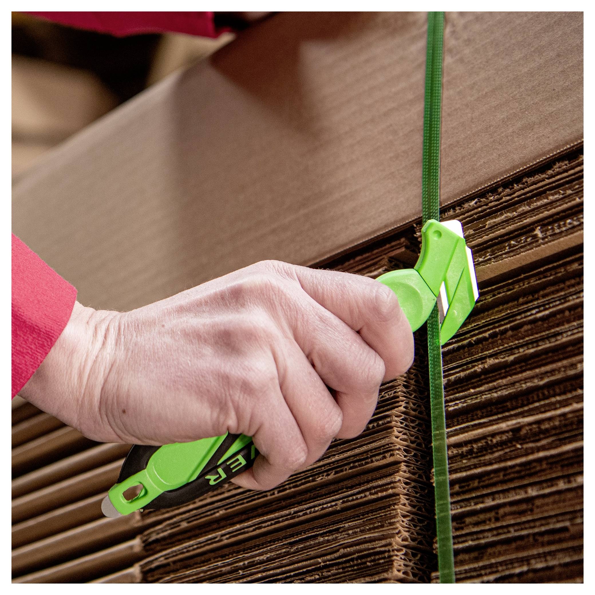Hand using a green strapping tensioner to tighten a plastic band around stacked cardboard sheets or boxes.