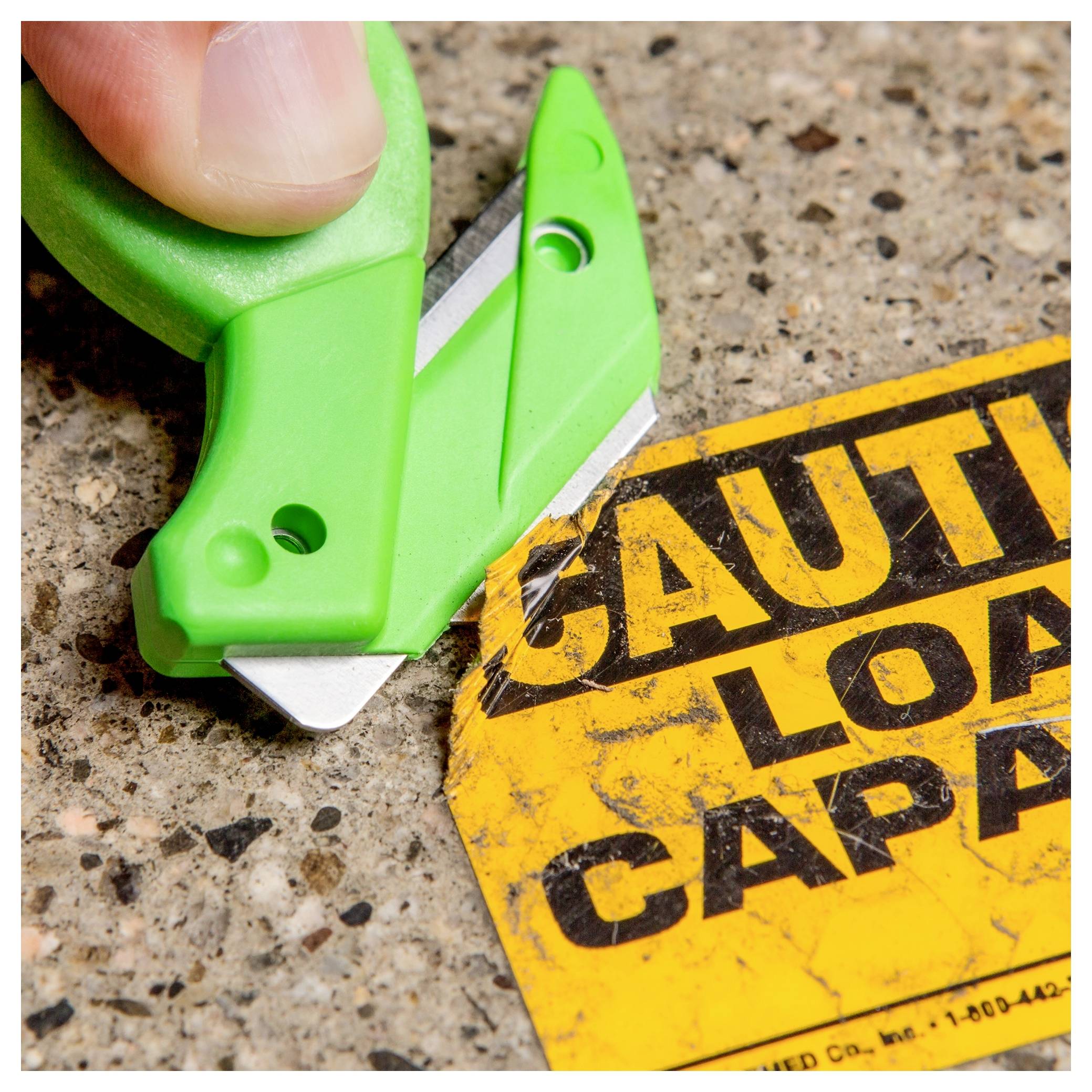 Green utility knife cutting a yellow 'CAUTION' label on a speckled surface, suggesting removal or scraping of adhesive signage.