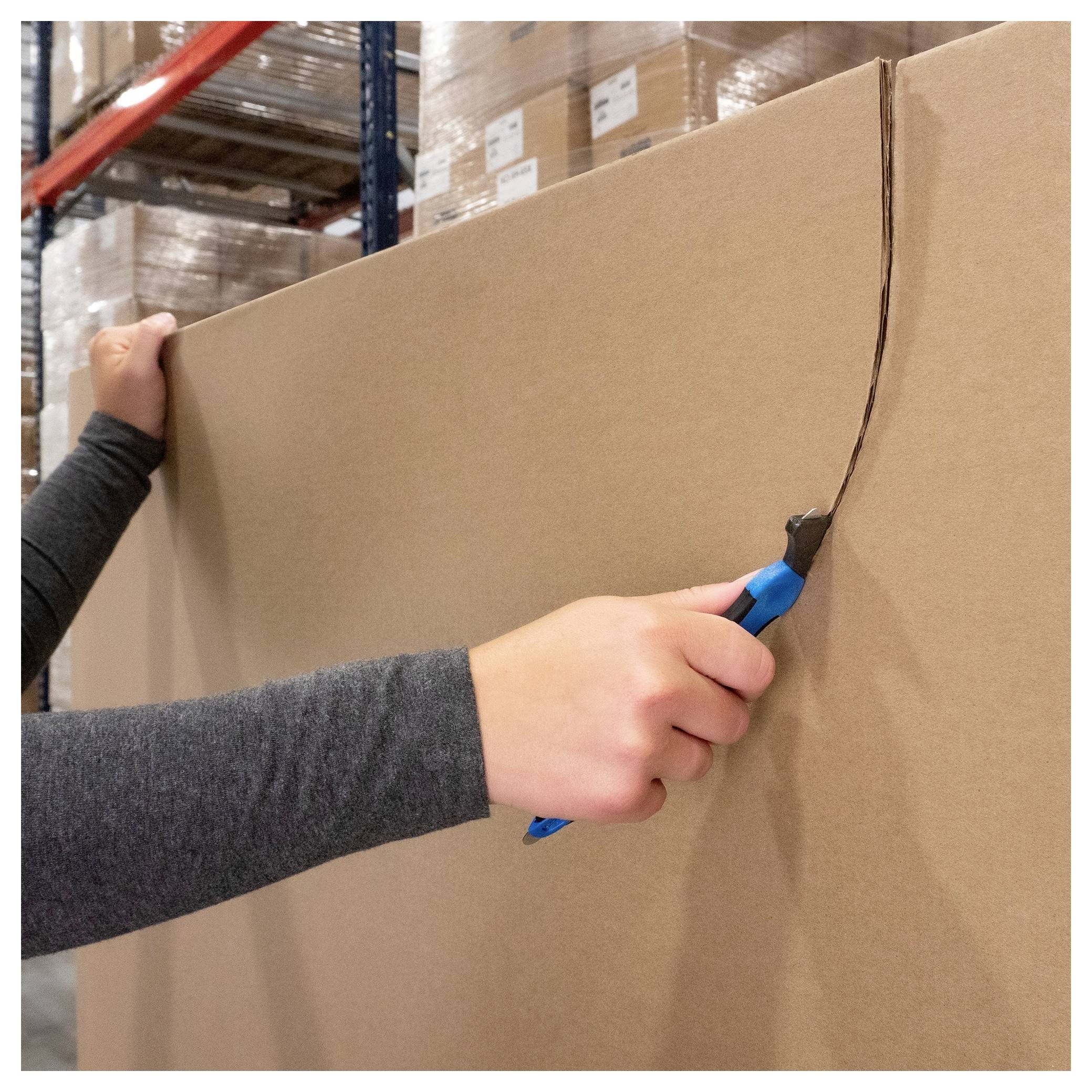 Person cuts open a large cardboard box with a utility knife in a warehouse, with stacked pallets in the background.