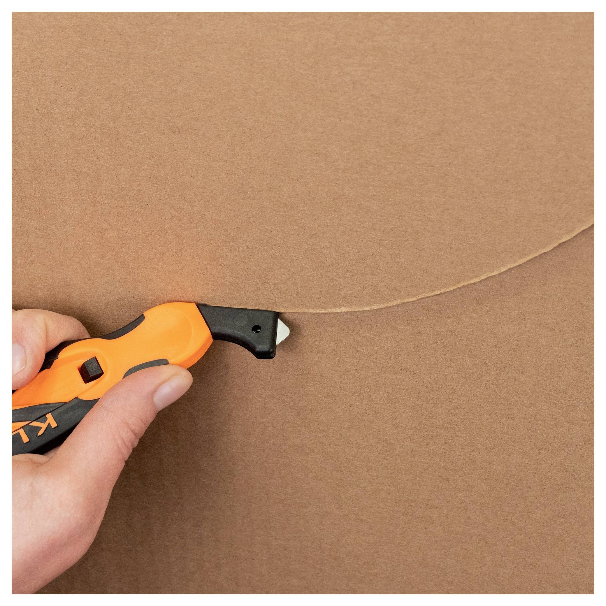 Hand using an orange utility knife to cut along a curved line on brown cardboard.