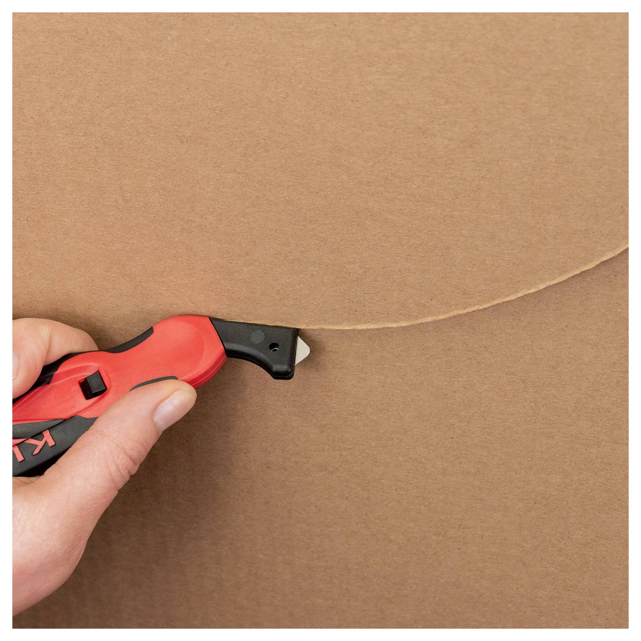 Hand using a red utility knife to cut a curved line into brown cardboard.<br>