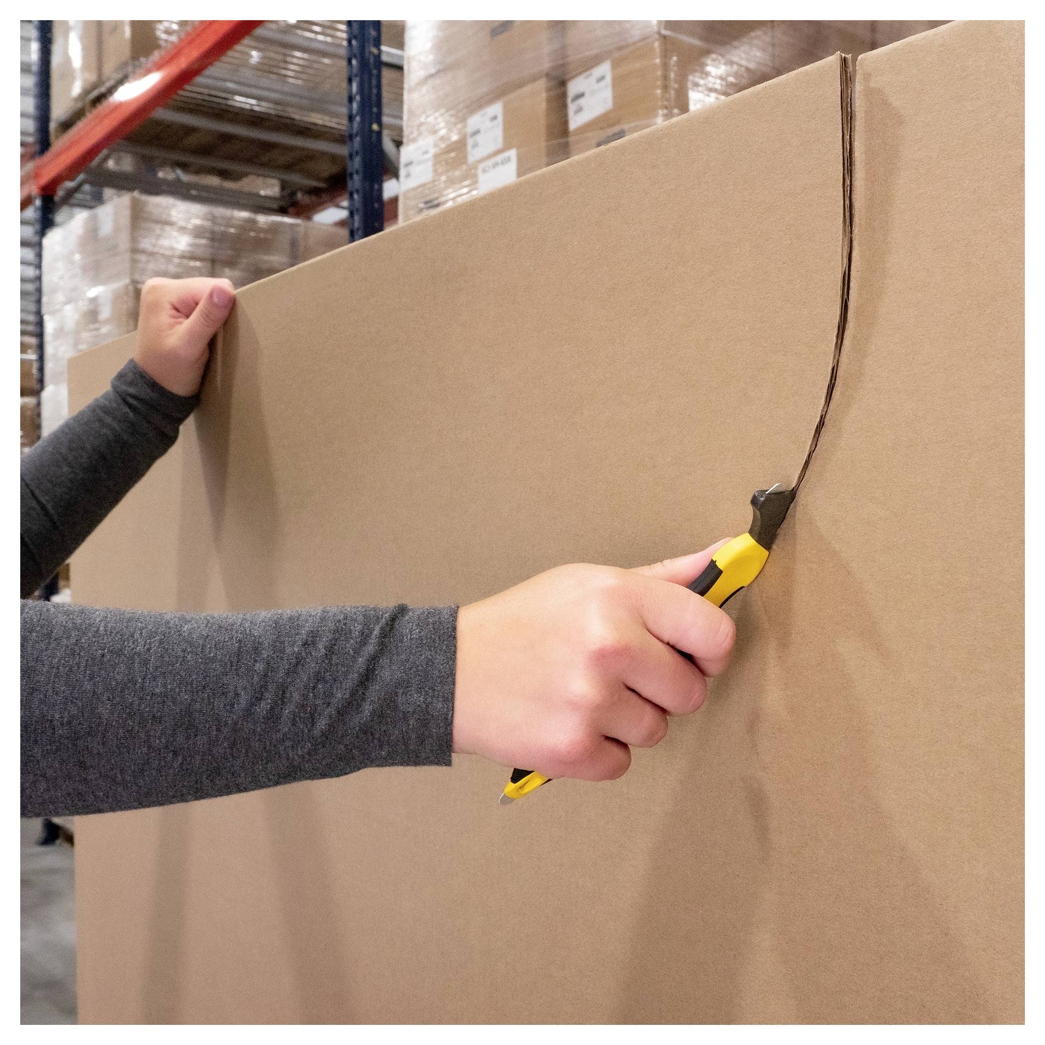 Person uses a utility knife to cut open a large cardboard box in a warehouse aisle.