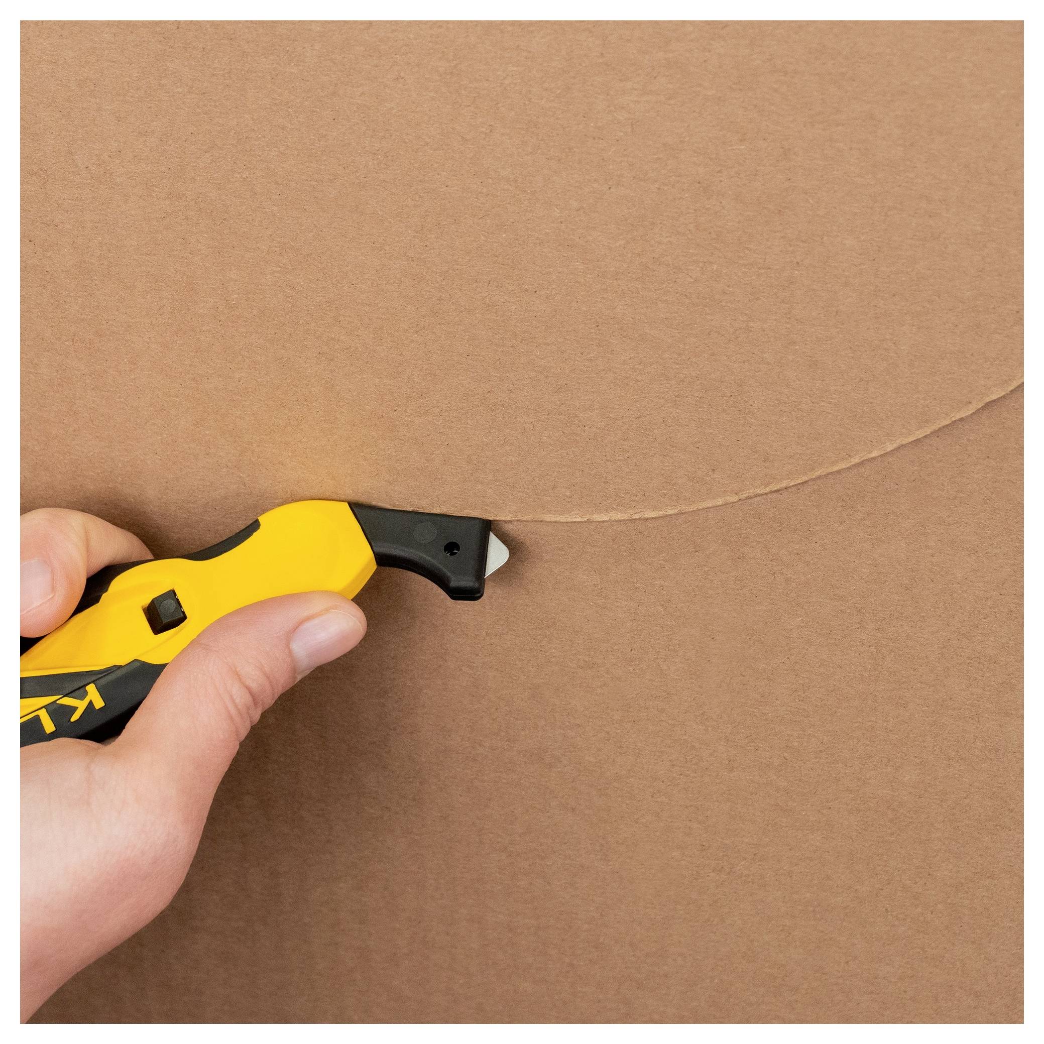 Hand using a yellow utility knife to cut through brown cardboard.