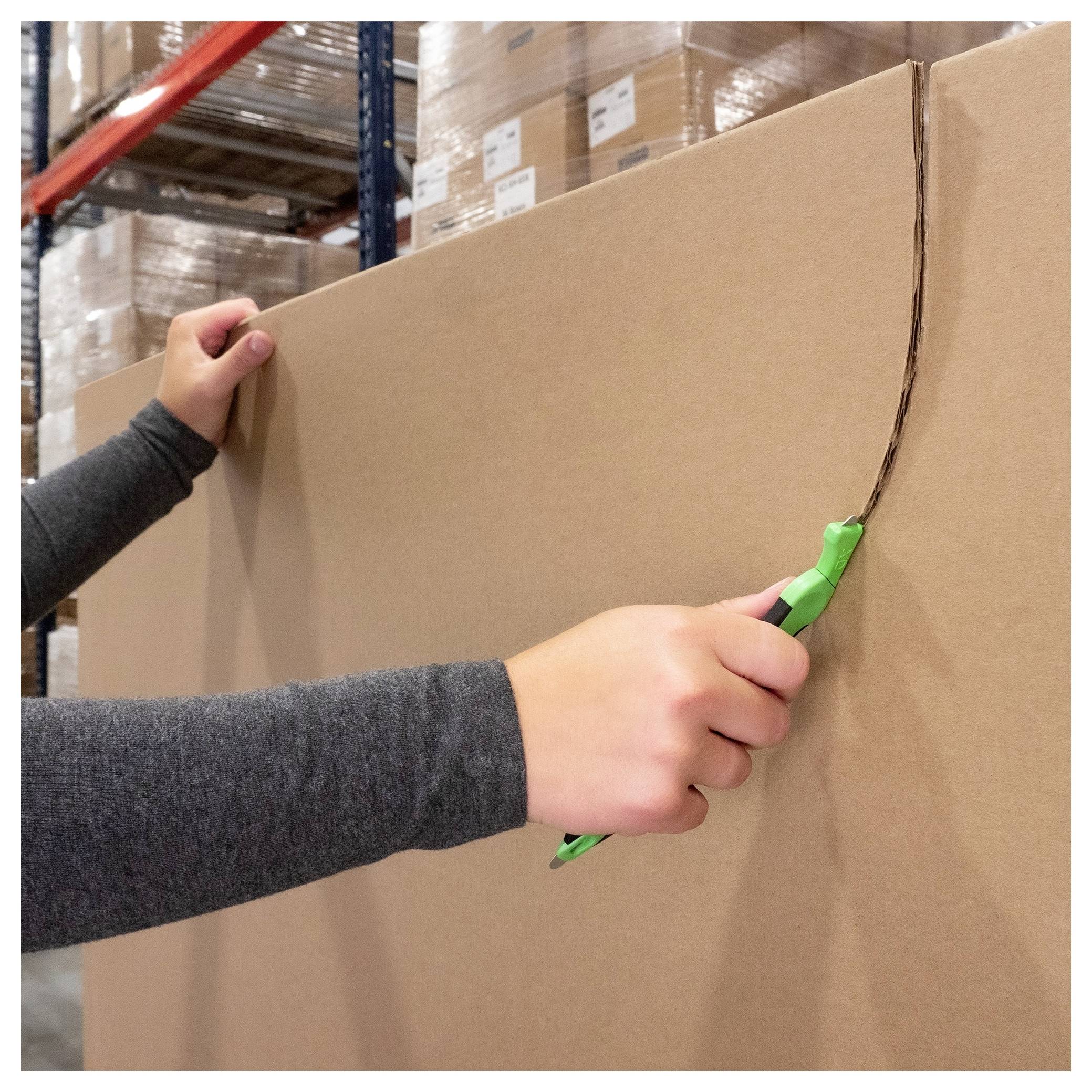 Hands using a utility knife to cut open a large cardboard box in a warehouse aisle with stacked pallets in the background.
