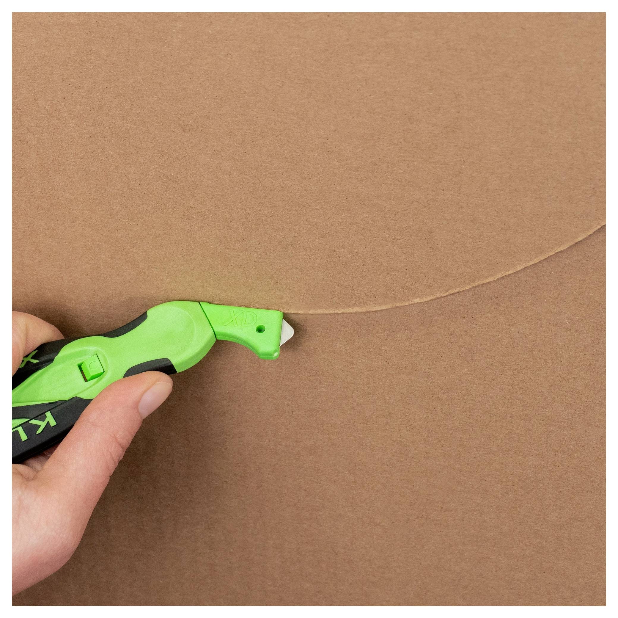 Hand holding a green utility knife cutting a curved line in brown cardboard.