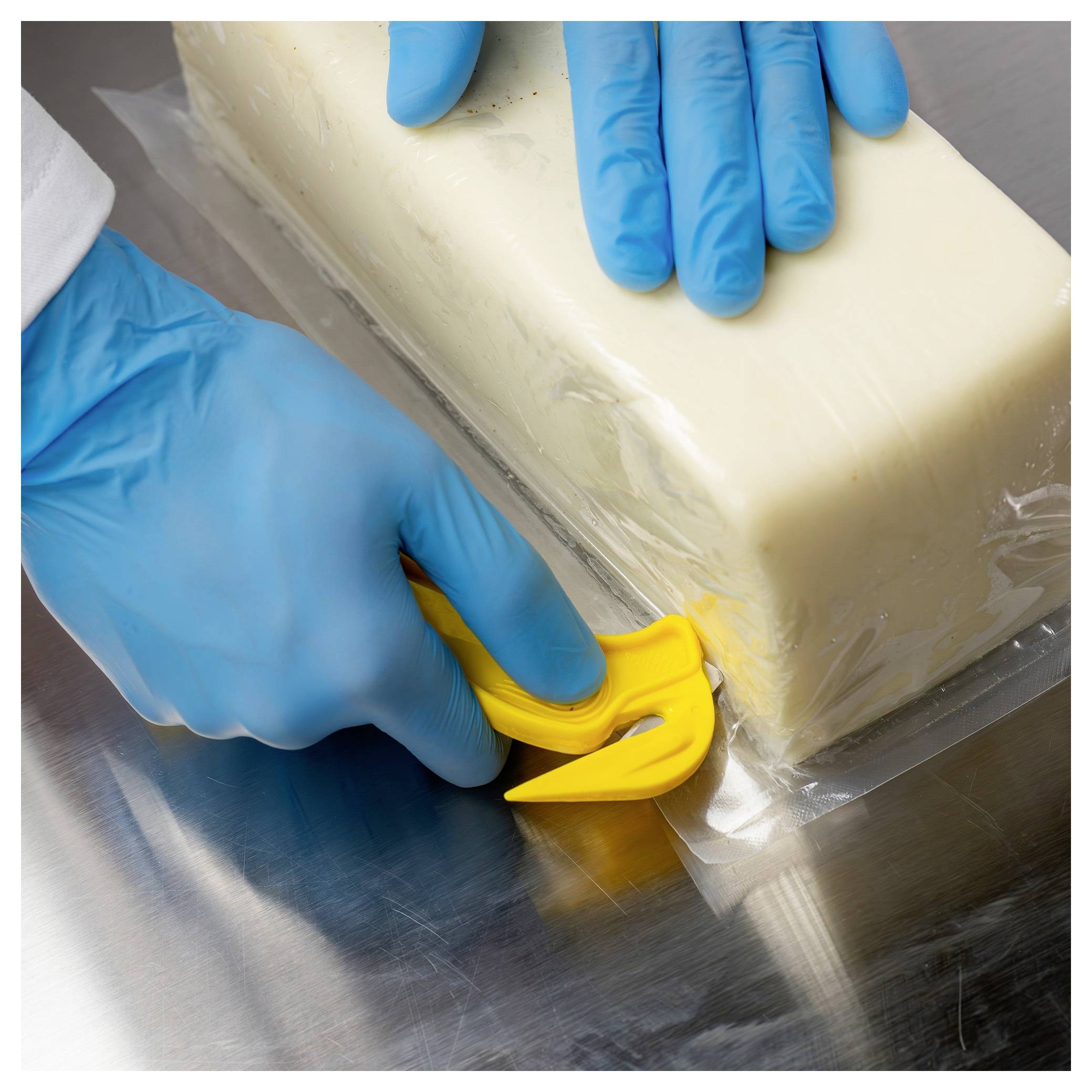 Gloved hands use a yellow cutter to open a wrapped block on a metal surface.<br>