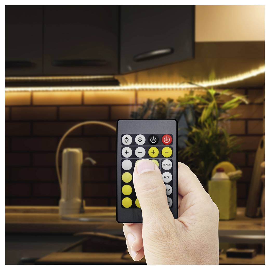 Hand holding a remote control aimed at an LED light strip in a kitchen, with the finger pressing a button to change the lighting color or brightness.