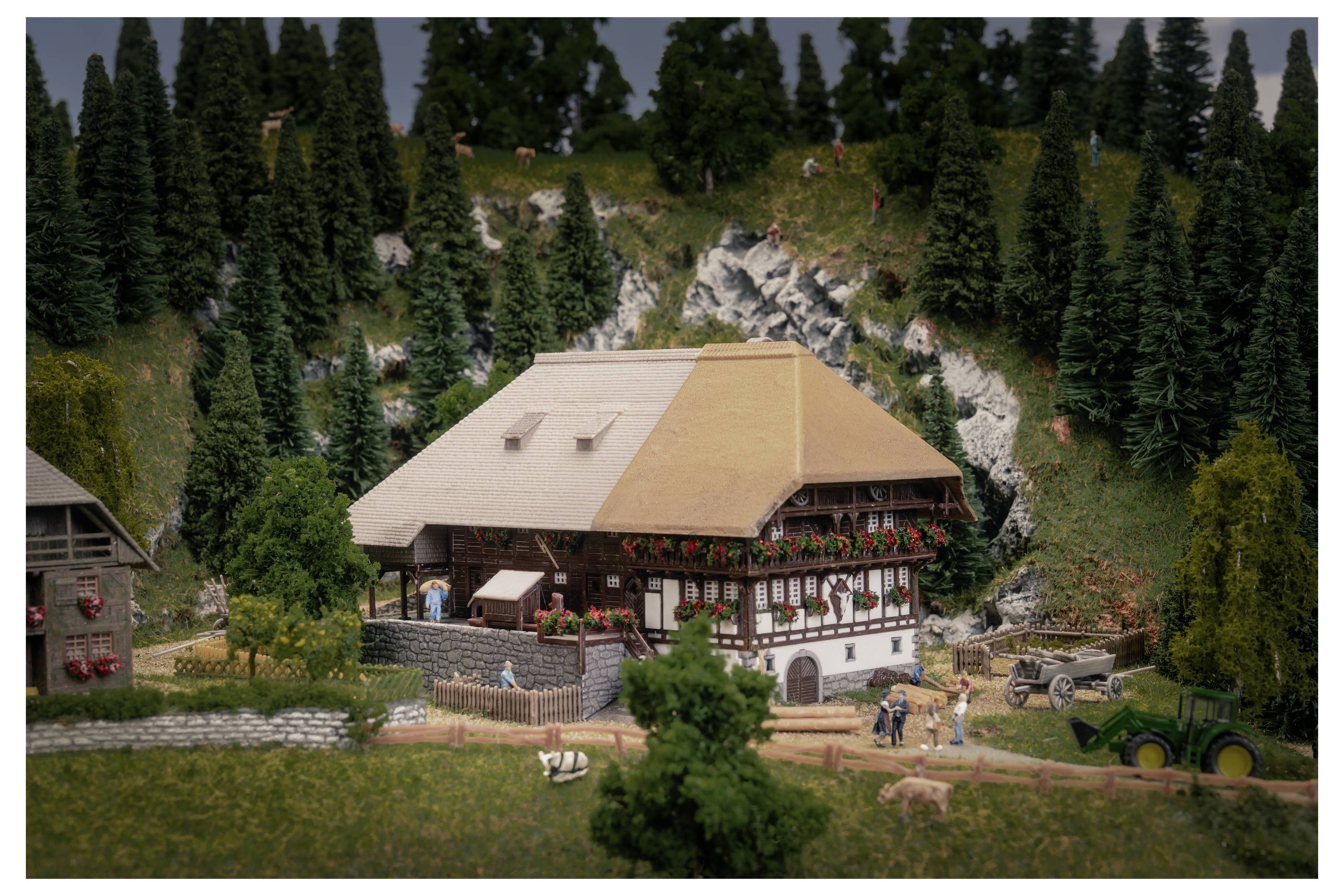 Model village scene with a large chalet-style house, people, small animals, and a tractor amid dense evergreen trees and rocky hillside.<br>