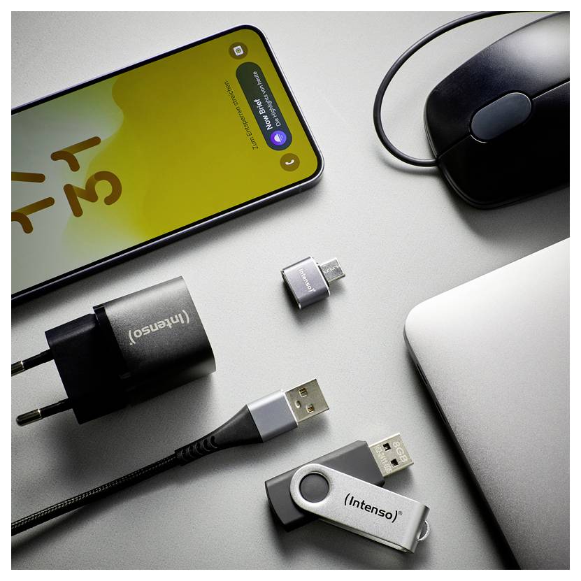 A smartphone, mouse, laptop corner, charger, and two USB adapters are arranged on a gray desk, with one adapter plugged into a cable.