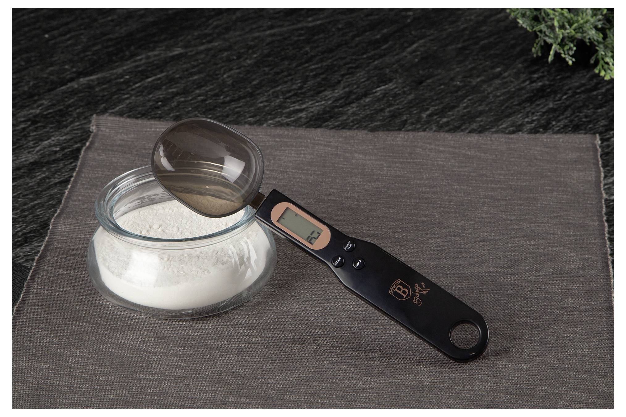 Digital kitchen scale spoon resting on a bowl of flour on a dark placemat.