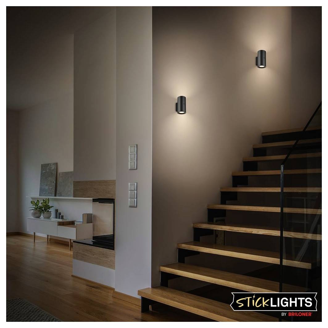 Modern staircase with two wall sconces casting light on a brown wall, beside an open living area with wood floors and a fireplace.