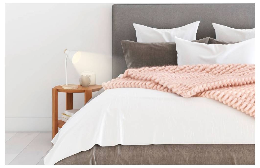 Neatly made bed with gray headboard, white bedding, pink knitted blanket, and a bedside table with lamp in a bright bedroom.