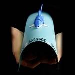 A light blue paper cone puppet with a small blue fish on top, held in two hands against a black background.
