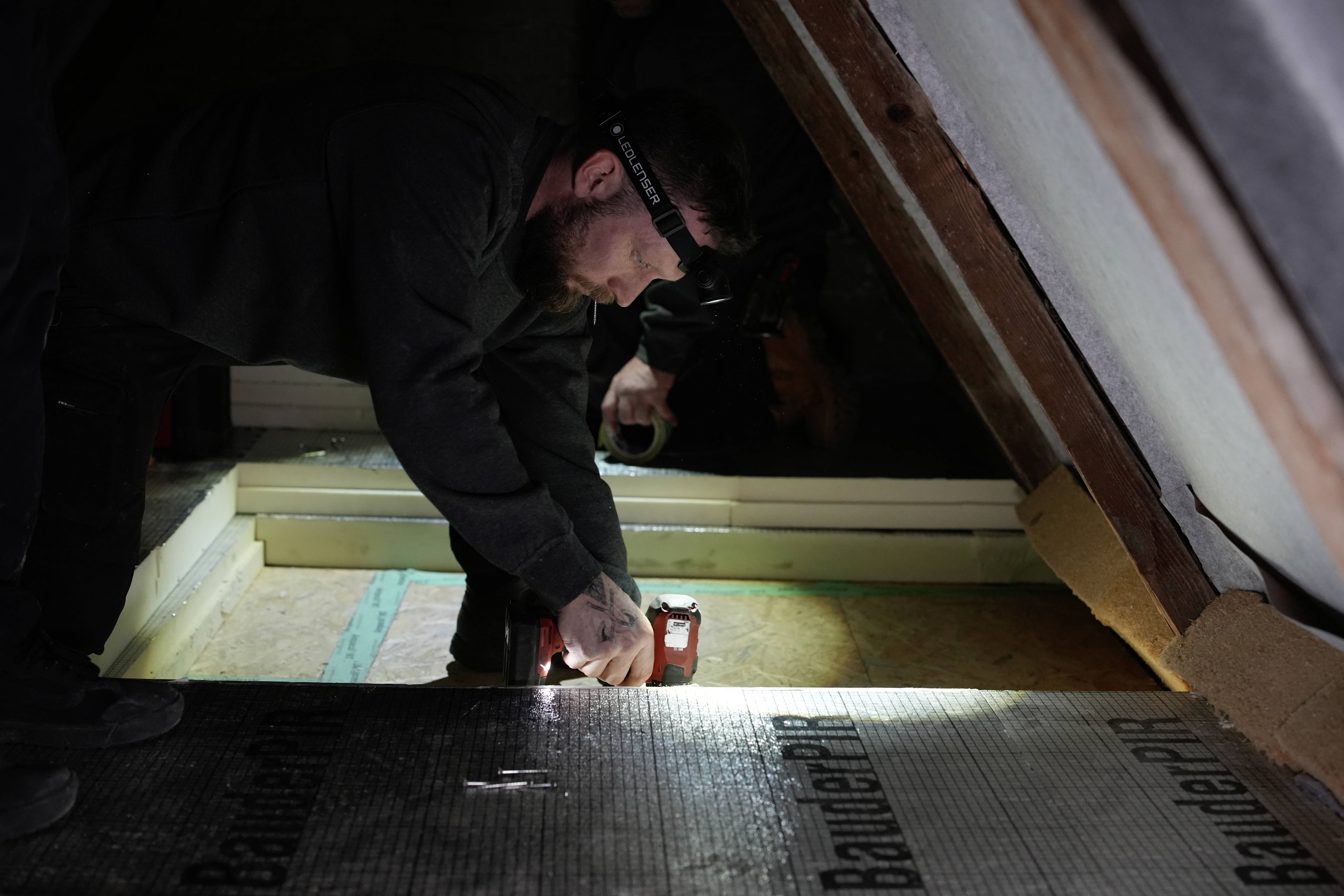 Worker in a dark attic uses a red power tool to fasten insulation boards between roof beams.