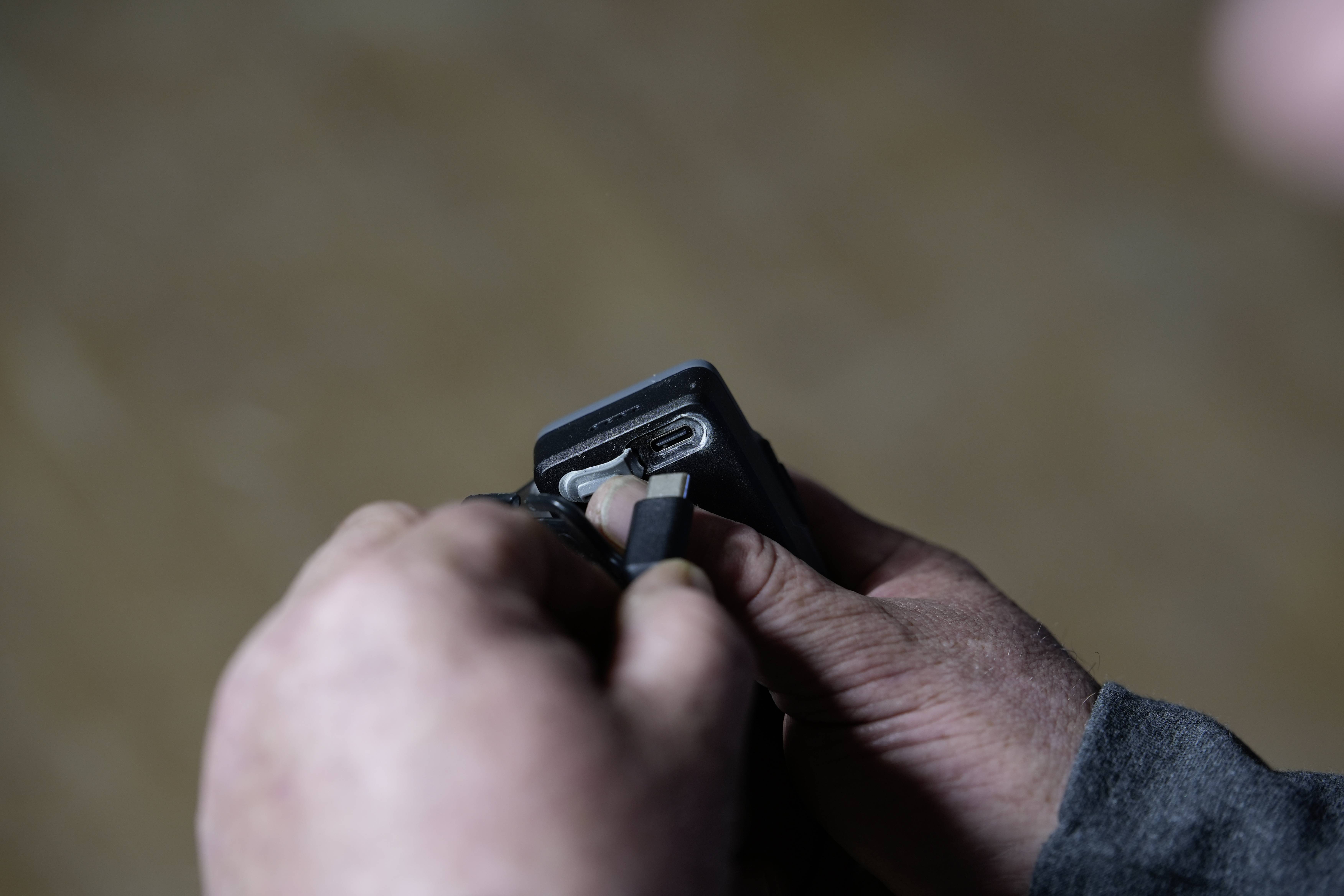 Close-up of hands plugging a black cable into a small device, with a blurred background.<br>