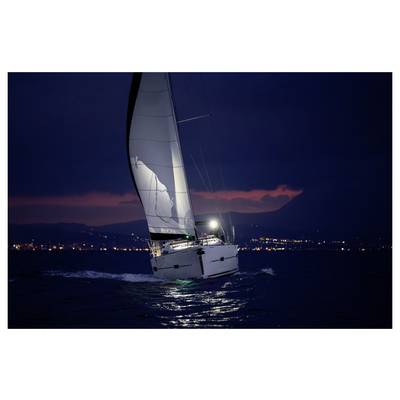 Sailboat with lit mainsail sailing at night on dark water, with distant city lights along the shore.