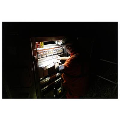 Worker in orange safety gear and helmet inspecting a brightly lit electrical control panel at night.<br>