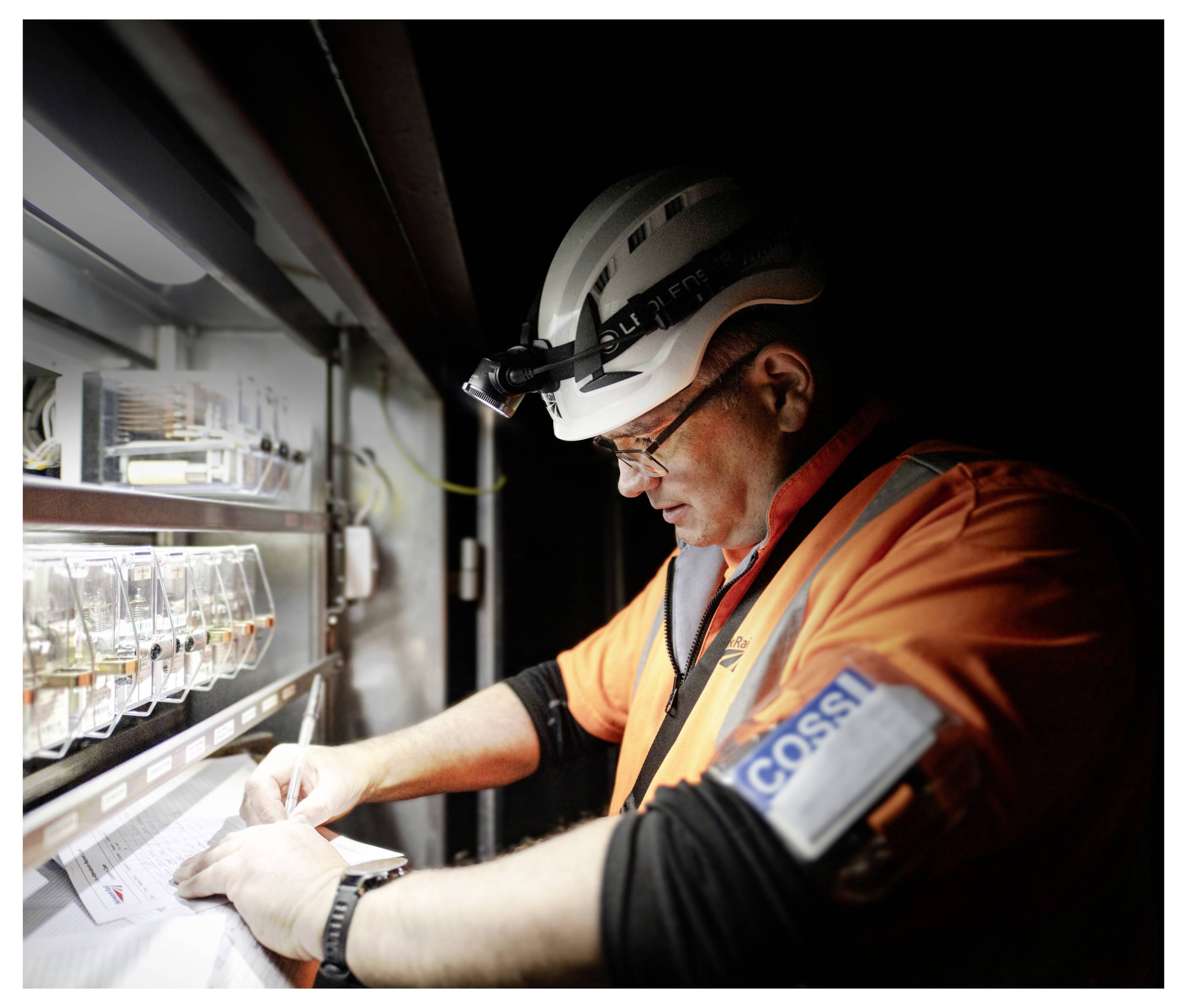Worker in hard hat and orange safety jacket records notes inside a dim industrial cabinet or inspection unit.