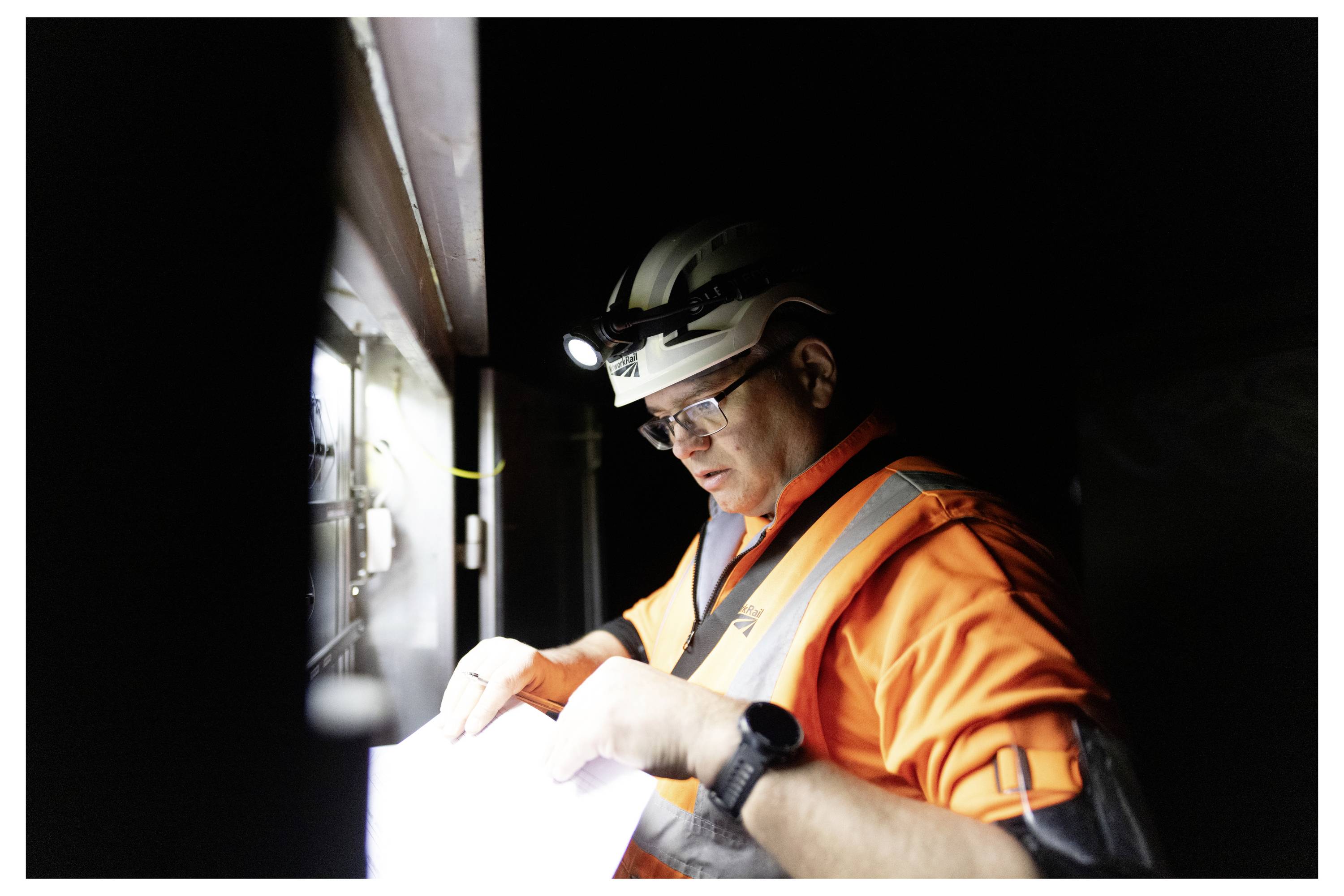 Construction worker in hard hat and safety glasses reviewing a document by flashlight in a dark work area.