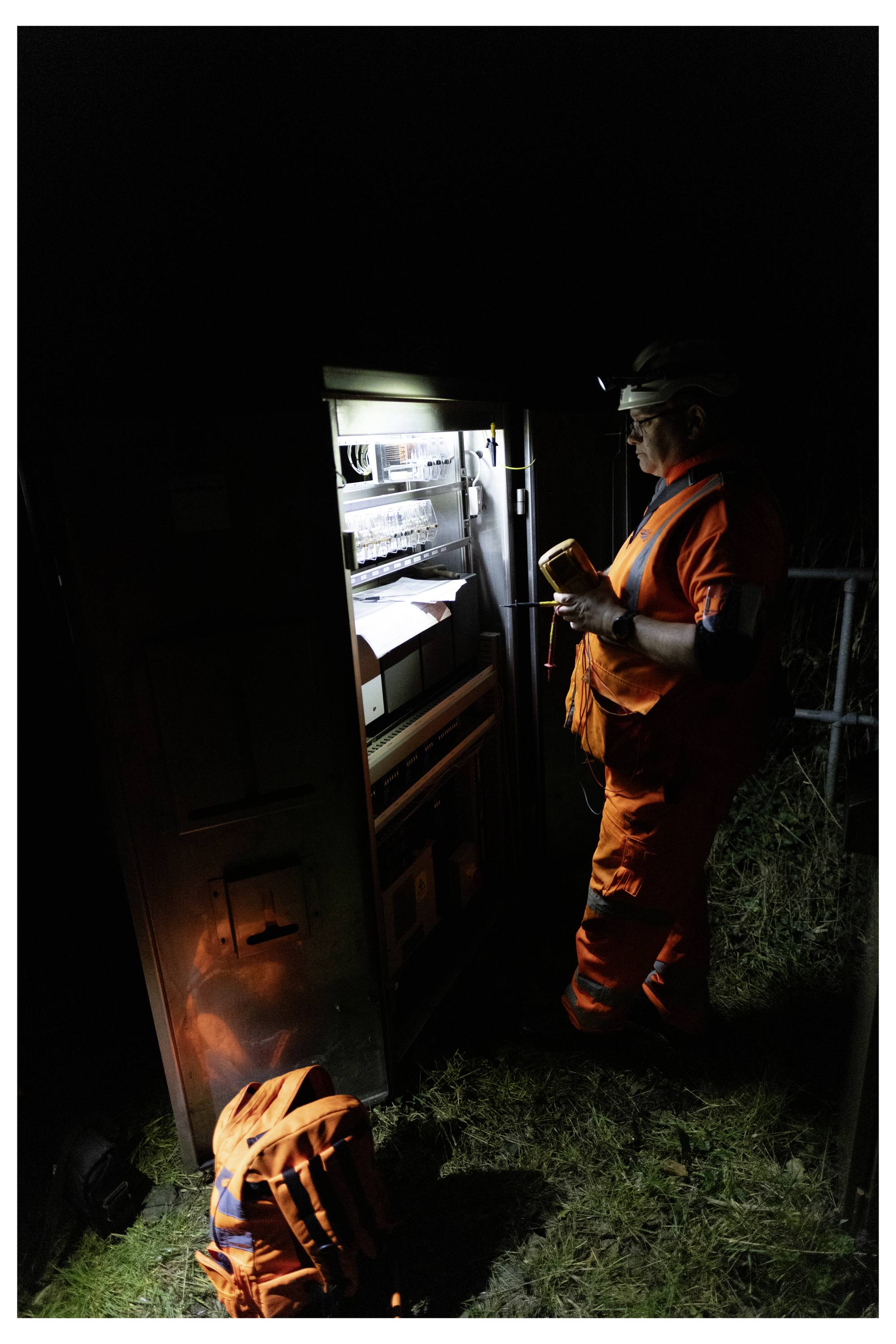 Worker in orange safety gear inspects equipment inside a lit roadside cabinet at night, with another orange bag or gear on the ground nearby.