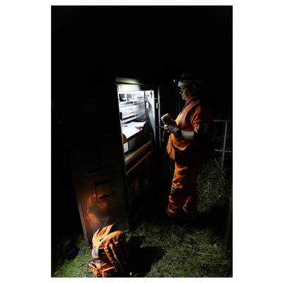 Worker in orange safety gear inspects equipment inside a lit roadside cabinet at night, with another orange bag or gear on the ground nearby.