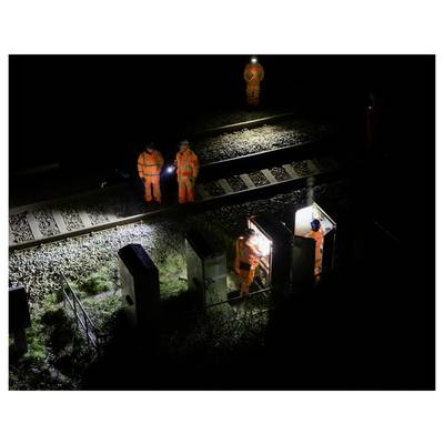 Workers in orange safety gear inspect a railway track at night, using a lit control box and flashlights.<br>