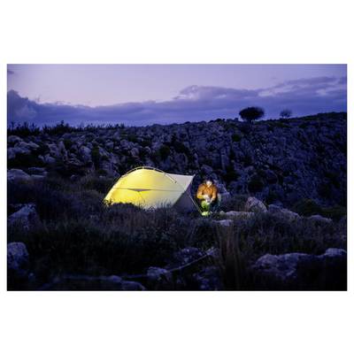 A person sits beside a glowing yellow tent at dusk on a rocky hillside, camping in a remote outdoor landscape.