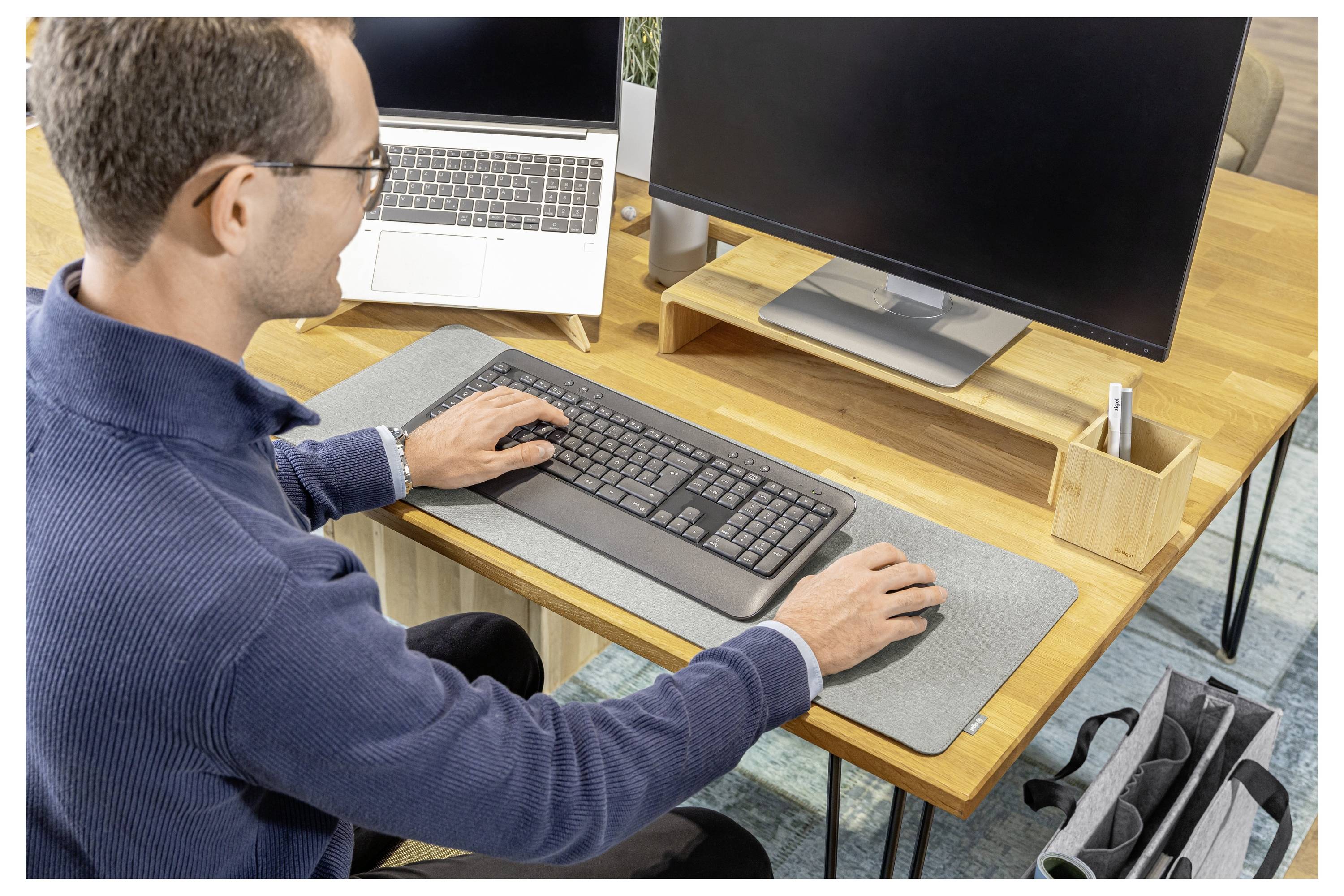 Man using a desktop computer at a wooden desk, typing on a keyboard beside a monitor and laptop in a home office.