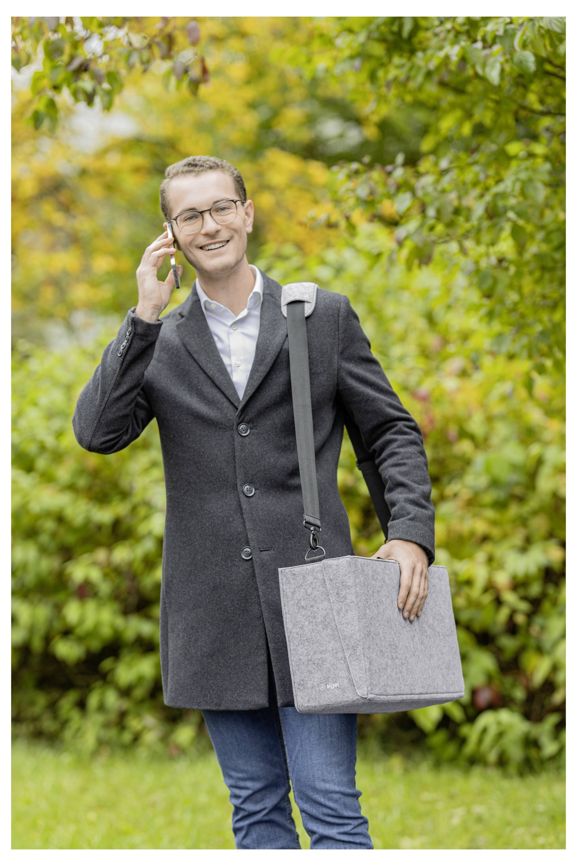 Smiling man in glasses stands outdoors in a park, talking on a phone while holding a gray laptop bag.<br>